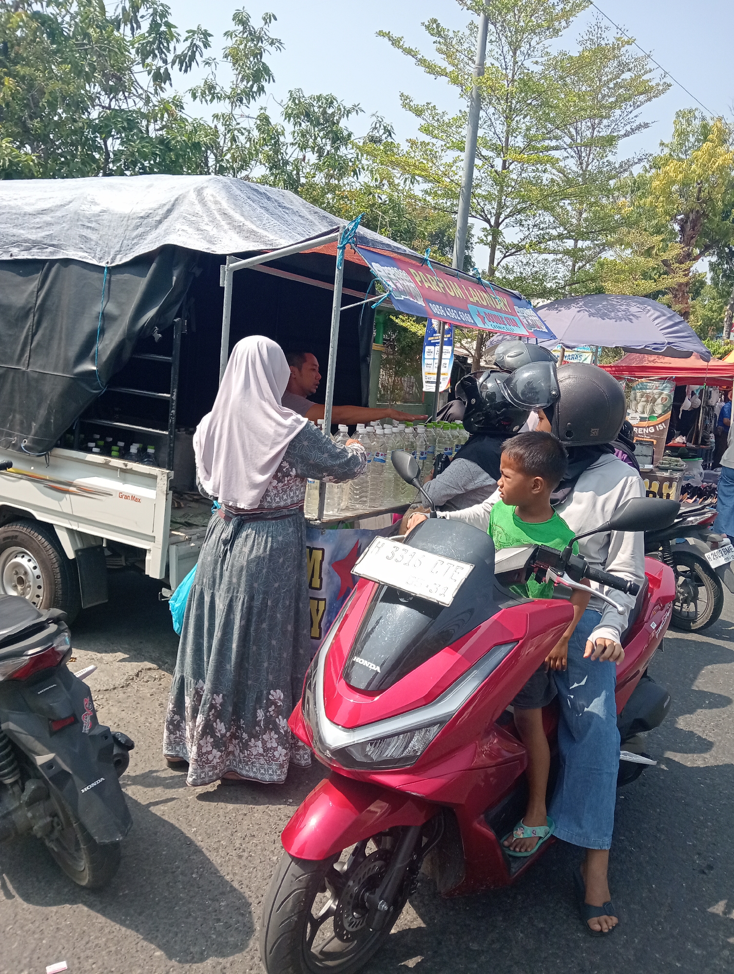 In Bintoro, Indonesia, on a sunny day, a woman wearing a light purple hijab and a long gray patterned dress is handing bottles of water to a man in the back of a white Gran Max truck. The truck is covered with a dark tarp. In the foreground, a red scooter is parked. On the scooter, a woman wearing a black helmet and a gray shirt is holding a young boy wearing a green t-shirt. The boy is looking to his left. The scooter has a license plate that reads "H 3315 GTE". In the background, there are trees, a street, and market stalls with colorful tarps.