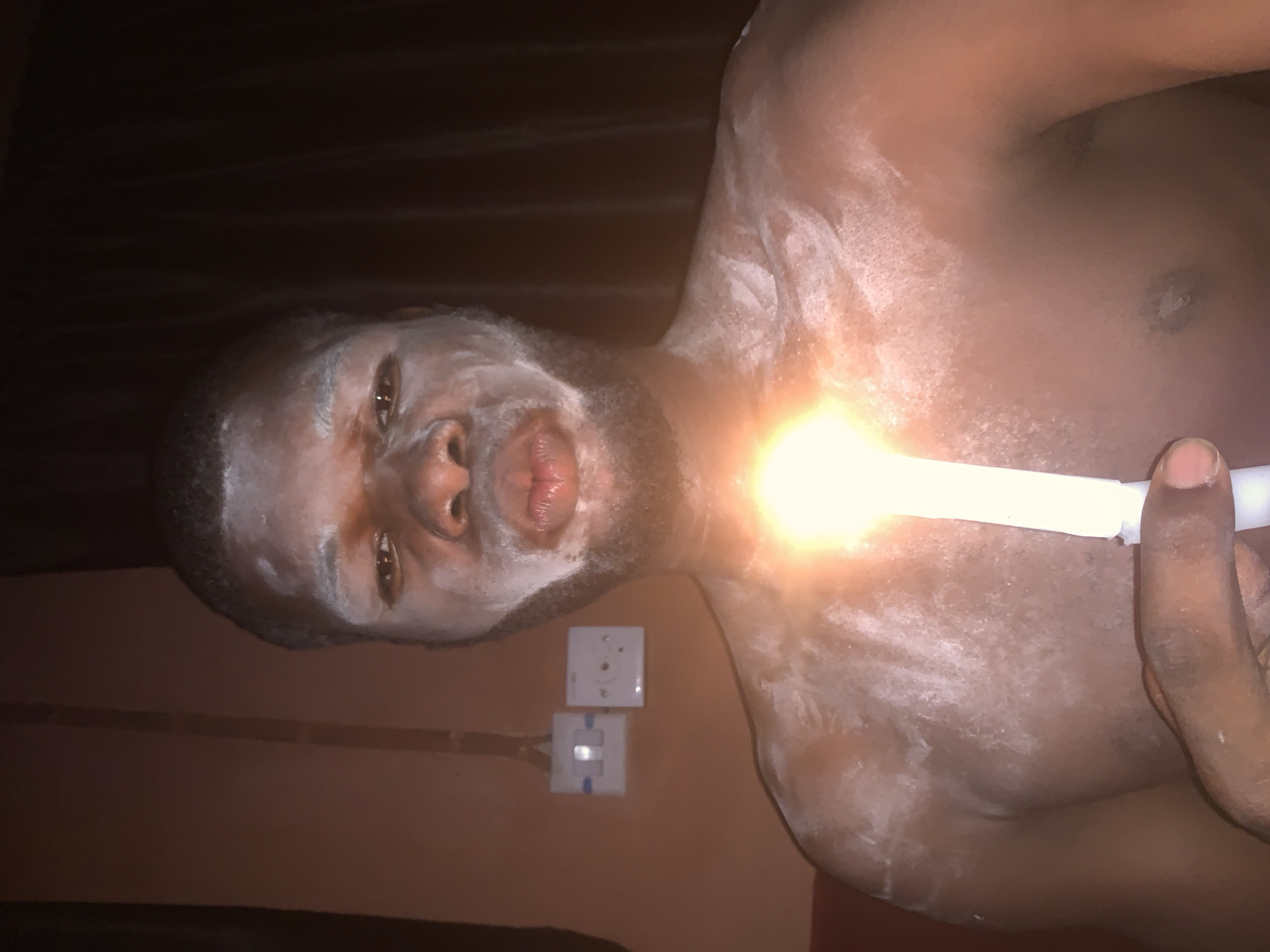 The image depicts a dark-skinned man in a dimly lit indoor setting, likely at night or in a room with minimal ambient light. His face, beard, neck, shoulders, and chest are generously covered with a white powdery substance. He looks directly at the viewer with a neutral expression, his mouth slightly open.
He holds a white, tube-like object in his right hand, which is emitting a very bright, intense yellowish-white light near his upper chest and neck area, creating a strong glare. The background features a reddish-brown wall where two white electrical outlets are visible. One appears to be a standard two-pin socket, and the other is a different type of white outlet. The style of the electrical outlets is consistent with those typically found in Nigeria, aligning with the given location context of Onitsha. The widespread application of the white powder on the man's body suggests a ritual, cultural practice, performance, or artistic endeavor.