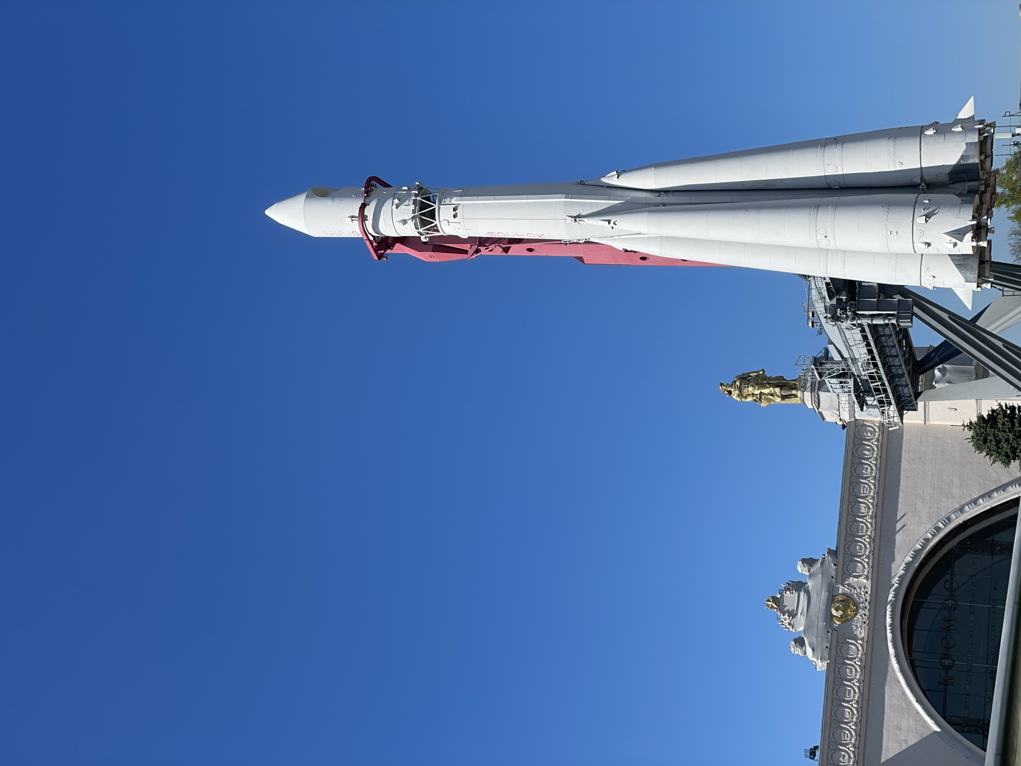 The image depicts a Vostok-class rocket on display in Moscow, Russia. The rocket, painted white with red accents, is mounted on a gray support structure. It appears to be a replica or a decommissioned vehicle. The background features a clear blue sky and part of a building with ornate decorations, including a gold statue. The building's architecture suggests it is part of the Exhibition of Achievements of National Economy (VDNKh) complex. The overall scene captures the grandeur of Soviet-era space achievements in a historical setting.