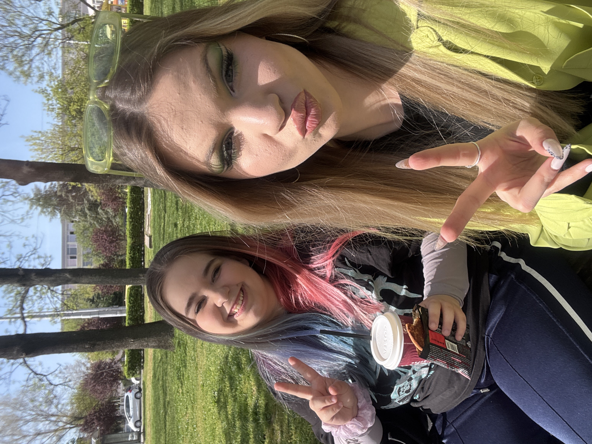This rotated image, likely a selfie, features two young women enjoying a bright, sunny day outdoors in what appears to be a park or green space in Plovdiv, Bulgaria.
The woman positioned at the top of the frame has long, straight blonde hair. She wears striking neon green rectangular sunglasses pushed up onto her head and a matching lime green jacket. Her makeup includes vivid green eyeshadow, and she is captured in a playful pose with pouting lips. Her left hand, adorned with white and black patterned nails and a silver ring on her middle finger, is raised.
The second woman, located at the bottom, has distinctive multi-colored hair in shades of pink, blue, and purple. She is smiling warmly and making a peace sign with her right hand, which has a light pink scrunchie on the wrist. She wears a dark top with a subtle light blue graphic, a light grey long-sleeved undershirt, and dark blue track pants with white side stripes. In her left hand, she holds a white "COSTA" branded coffee cup and a dark-colored snack bar wrapper.
The background reveals a sunny environment with clear blue skies, vibrant green grass, and various trees – some displaying fresh spring leaves, others with reddish foliage – and hedges. In the distance, parts of a building and a white car are visible. The overall scene conveys a cheerful and relaxed atmosphere, suggesting a pleasant leisure time for the two friends.