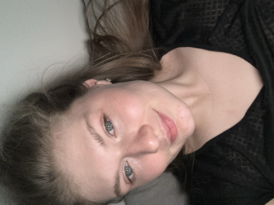 A woman lies on a surface with her head tilted towards the camera. She has long brown hair and blue eyes. She is wearing a black patterned top. Her skin appears fair with some visible texture and minor blemishes. A small earring is visible in her left ear. The background is a light, neutral color. She is smiling gently.