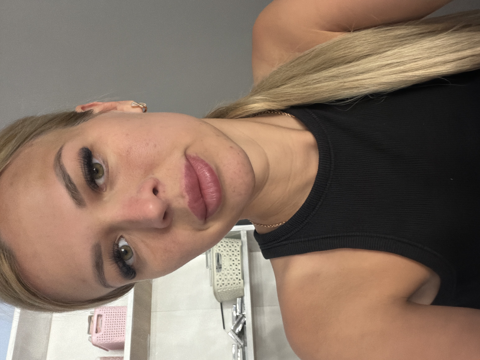 The media file is a portrait of a young woman with blonde hair, wearing a black tank top and a gold necklace. She is looking at the camera with a neutral expression. Her hair is pulled back into a ponytail, and she is wearing makeup, including false eyelashes and lip gloss. There is a ring on her left ear. In the background, there are some shelves with decorative baskets and what appear to be toiletries. The lighting suggests an indoor setting, possibly a bathroom or dressing room. The image appears to be taken with a smartphone, as indicated by the close-up framing and the upward angle. The setting is likely domestic, given the presence of personal items and the relaxed pose of the subject. There is no visible text in the image.