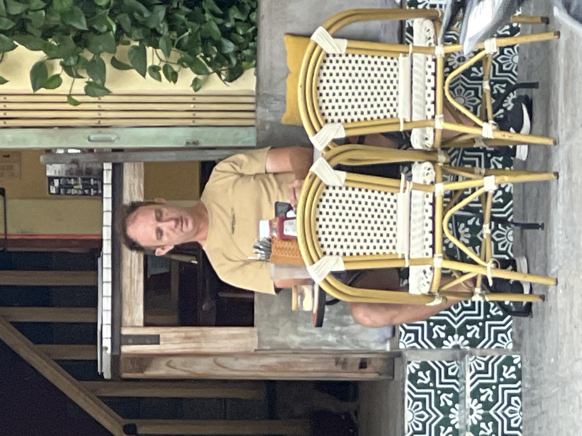A man is seated at a table outside a building. He is wearing a tan t-shirt and shorts. Two chairs with yellow frames and woven seats are positioned in front of the table. A basket of utensils and two bottles of condiments are on the table. The building has wooden and grey elements, and there is greenery to the right. The ground in front of the building is tiled with a green and white geometric pattern. The scene appears to be in Nha Trang, Vietnam, as indicated by the presence of Vietnamese text on a menu visible on the table, and the general ambiance of a street-side cafe.