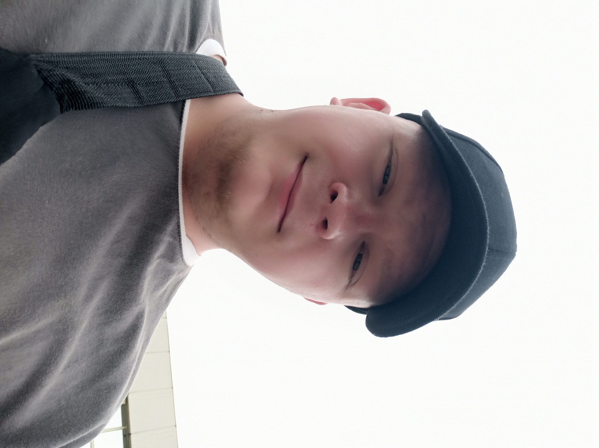 An outdoor, eye-level, waist-up, medium shot shows a fair-skinned, bald man wearing a black baseball cap and a gray t-shirt. The man is looking directly at the camera and smiling slightly, with his head tilted to his left. A black strap is visible across his chest from the t-shirt. The background is a bright, overcast sky, with a sliver of a building visible on the right side of the frame. The man's face and upper body occupy most of the frame, with the background appearing out of focus.