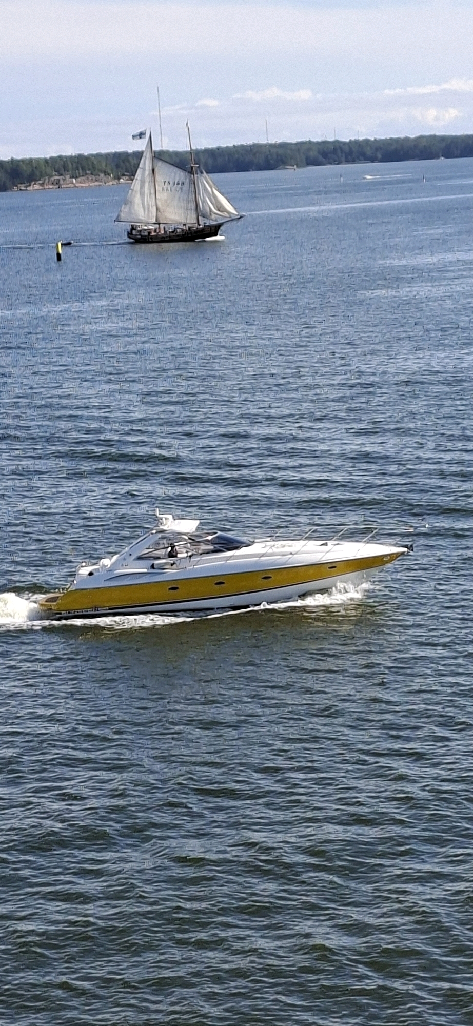 In Helsinki, Finland, two boats are depicted on the water. In the foreground, a white and yellow motor yacht with a closed cabin speeds through the water, leaving a wake. The yacht has a dark tinted windshield and chrome railings. In the background, a traditional sailing ship with two masts and white sails moves slowly through the water. The ship appears to be a historic vessel. The water is blue with moderate waves. A line of green trees forms the background on the distant shore. The sky is light blue with scattered white clouds. A yellow buoy is visible to the left of the sailing ship.