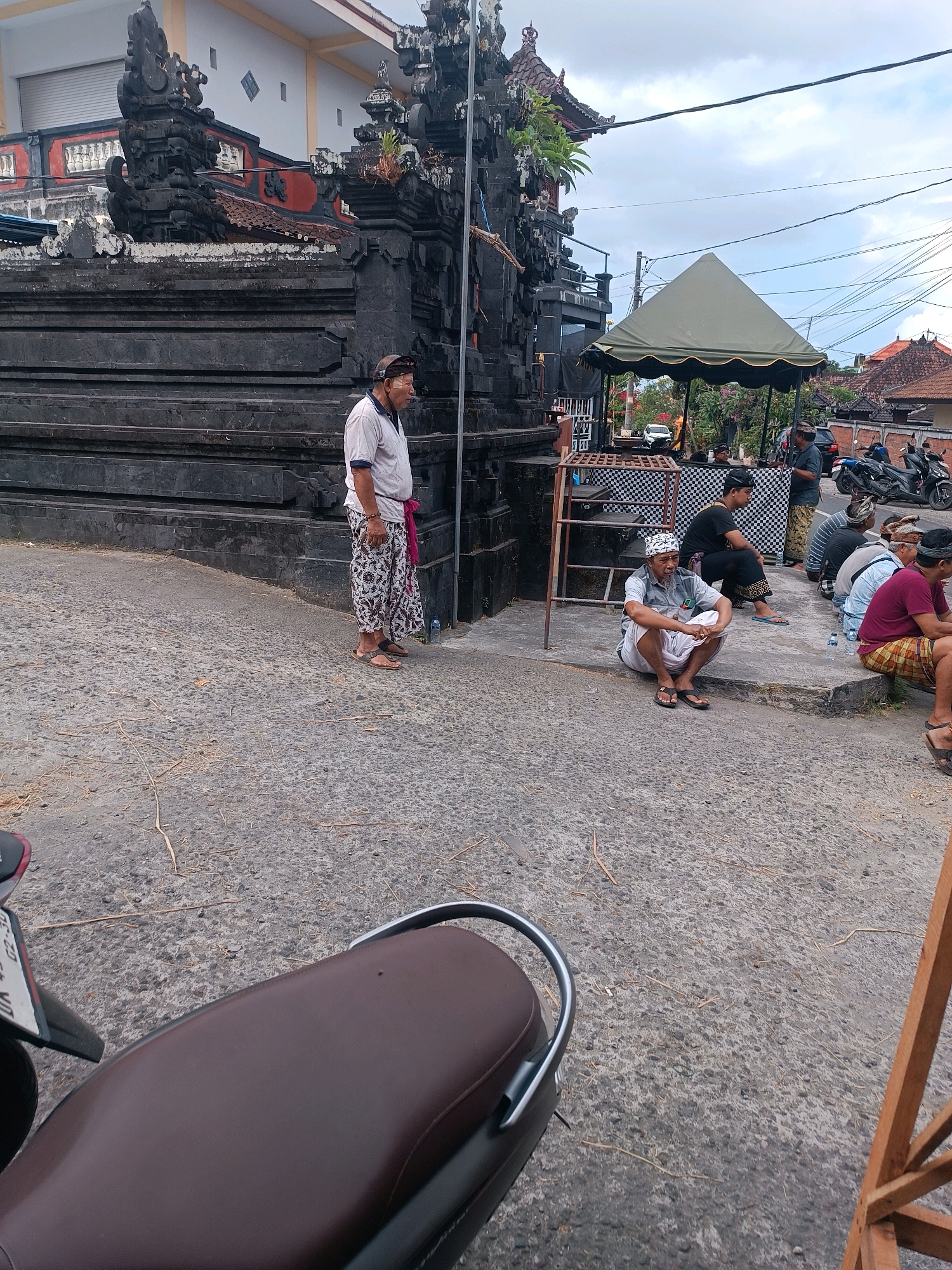 In Selemadeg, Indonesia, a group of men are gathered near a traditional Balinese temple entrance. A man in a white shirt and patterned pants stands to the left, facing away from the camera. To the right, several men are seated on the ground. One man, wearing a white headdress, sits cross-legged in the foreground, facing the camera. Behind him, other men sit in a row, some wearing traditional Balinese attire. A large, dark stone structure, likely part of the temple, dominates the left side of the image. A green tent is set up in the background, with motorcycles parked nearby. The ground is paved with asphalt and scattered with dry leaves and twigs. The sky is overcast.