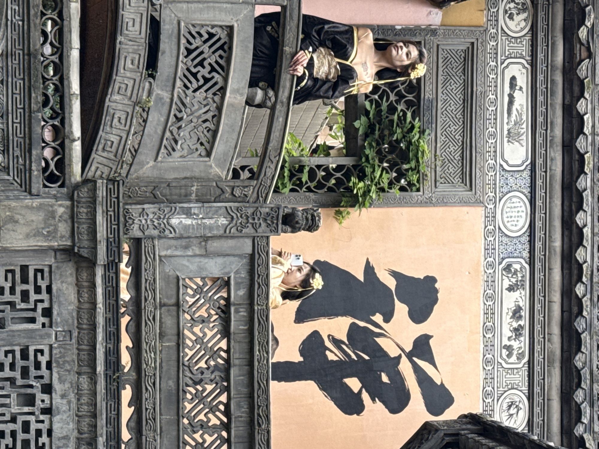 A woman wearing a black and gold dress is leaning out of a building window with ornate gray stone latticework. Another woman, also wearing a yellow flower in her hair, is seen in another window on the right taking a picture with her cellphone. The building has peach-colored walls with black traditional Chinese calligraphy on the side. The building's style is that of Yuzhong District, China.