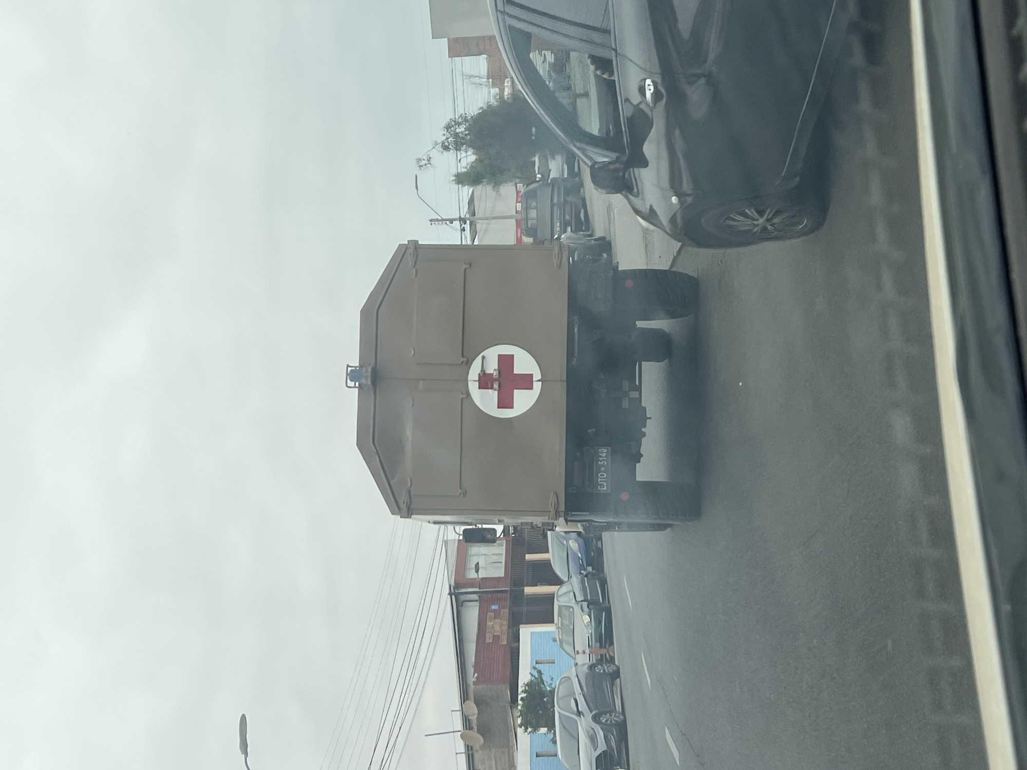 The image captures a street scene in Arica, Chile, viewed from inside a vehicle, looking out the passenger side window. The primary subject is a large, olive-drab or tan colored military-style truck, possibly a medical transport or ambulance, seen from the rear. A prominent red cross symbol on a white circular background is affixed to the back of the truck, indicating its medical purpose.
The truck is equipped with a license plate that reads "CJTO - 5140". "CJTO" likely stands for "Ejército," meaning Army in Spanish, confirming it as a military vehicle. It appears to be traveling on a paved road, possibly in traffic, as other vehicles are visible both in front and behind it in adjacent lanes. A dark grey sedan is partially visible to the right, and other cars, including a blue and a lighter grey vehicle, are further down the road.
The setting is an urban or semi-urban environment, characterized by various buildings lining the street, power lines, and utility poles with a street light. Sparse green foliage or trees can be seen in the background. The sky is overcast and light, suggesting daytime with cloudy weather. No specific activity or event beyond general road travel is discernible. There are no clearly visible individuals in the image.