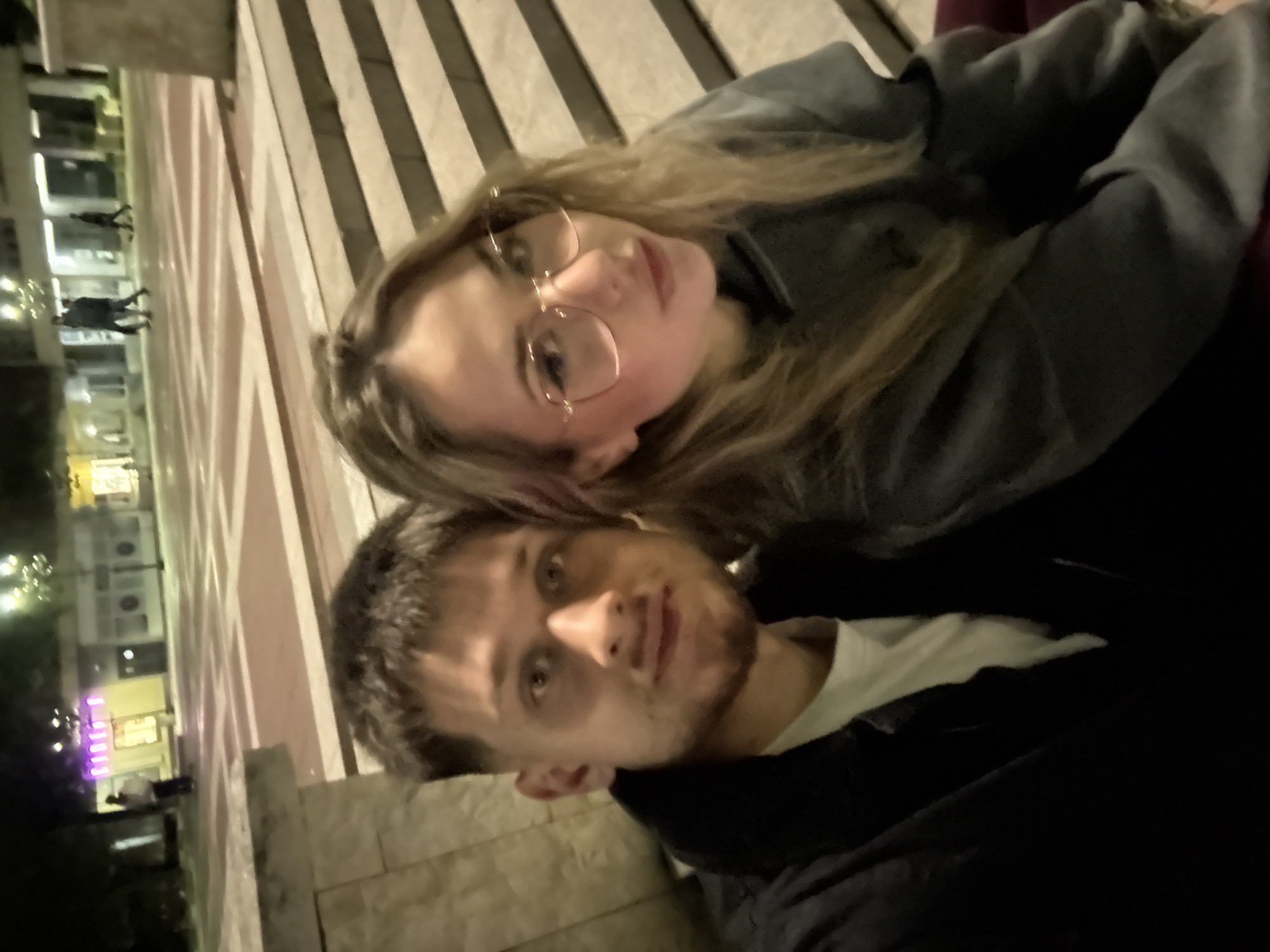 Two people are posing for a selfie, possibly in Blagoevgrad, Bulgaria. The woman is fair-skinned, with blonde hair, and is wearing glasses. The man has short, dark hair and a trimmed beard. They are positioned in front of a background of striped pavement, with a building visible in the distance. The picture appears to have been taken at night, given the lighting conditions.