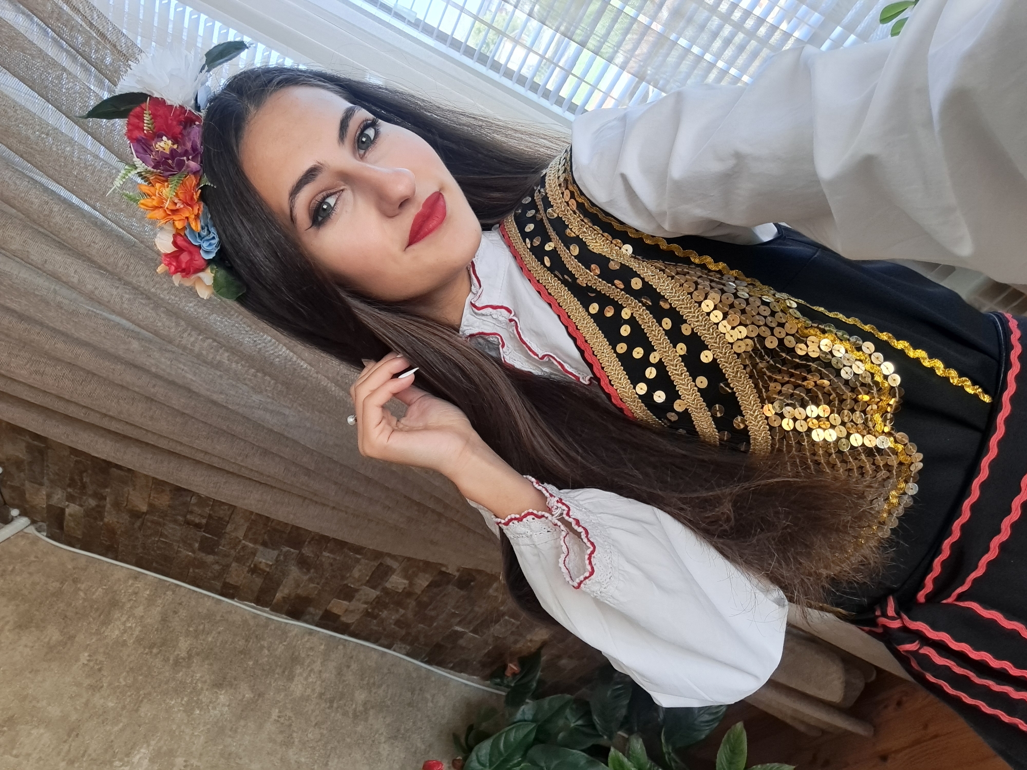A young woman in traditional Bulgarian folk costume is photographed indoors, seemingly in Unknown, Unknown. She has long, straight brown hair, red lipstick, and a floral headpiece. She wears a white blouse with lace-trimmed sleeves and a black vest adorned with gold sequins and red trim. The background consists of a window with sheer curtains and a partially visible tiled wall. A potted plant is visible in the lower left corner. The overall shot appears to be a selfie.