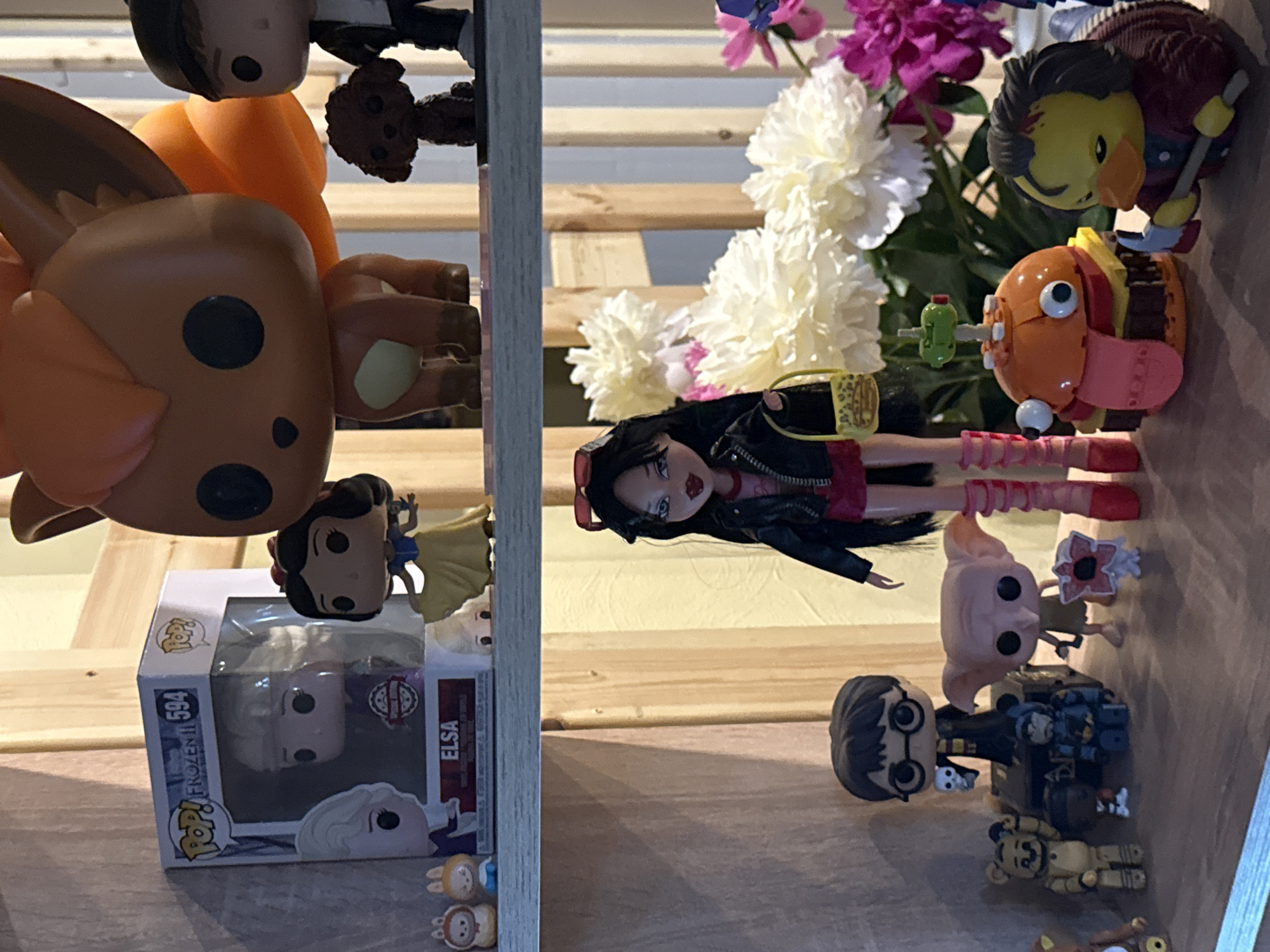 This is a medium shot, eye-level, indoor photograph of a collection of figurines and collectibles displayed on wooden shelves. The background is composed of vertical wooden planks.
On the top shelf, from left to right, there are two small bunny figurines, a Funko Pop! box containing an Elsa figurine from "Frozen II," a Snow White Funko Pop! figurine, a large orange and brown fox-like creature Funko Pop! figurine, a small dark brown poodle figurine, and a Funko Pop! figurine of a person wearing a black jacket.
On the bottom shelf, from left to right, are a small yellow bear figurine, a black cube-shaped object, a Harry Potter Funko Pop! figurine holding a skull, a small blue and black figurine, a Dobby Funko Pop! figurine, a small red and white flower-like figurine, a doll with long black hair, red sunglasses, a black leather jacket, and pink boots, holding a yellow purse, a burger-shaped toy with googly eyes and a tongue, and a yellow duck figurine dressed as a character from "The Shining" holding an axe. Behind the figurines, there are large white and pink artificial flowers. The lighting appears to be artificial and warm.
The image does not contain any information indicating a location in Khimki, Russia.