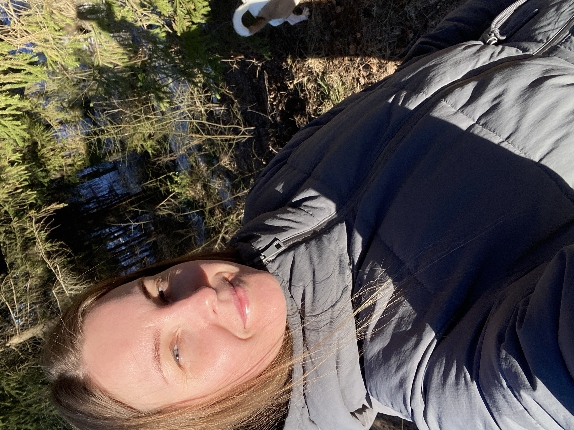 A woman wearing a gray padded jacket is lying on her back in an area with trees and some remaining snow. Part of a white dog is visible in the background. The location is Noviy, Russia.