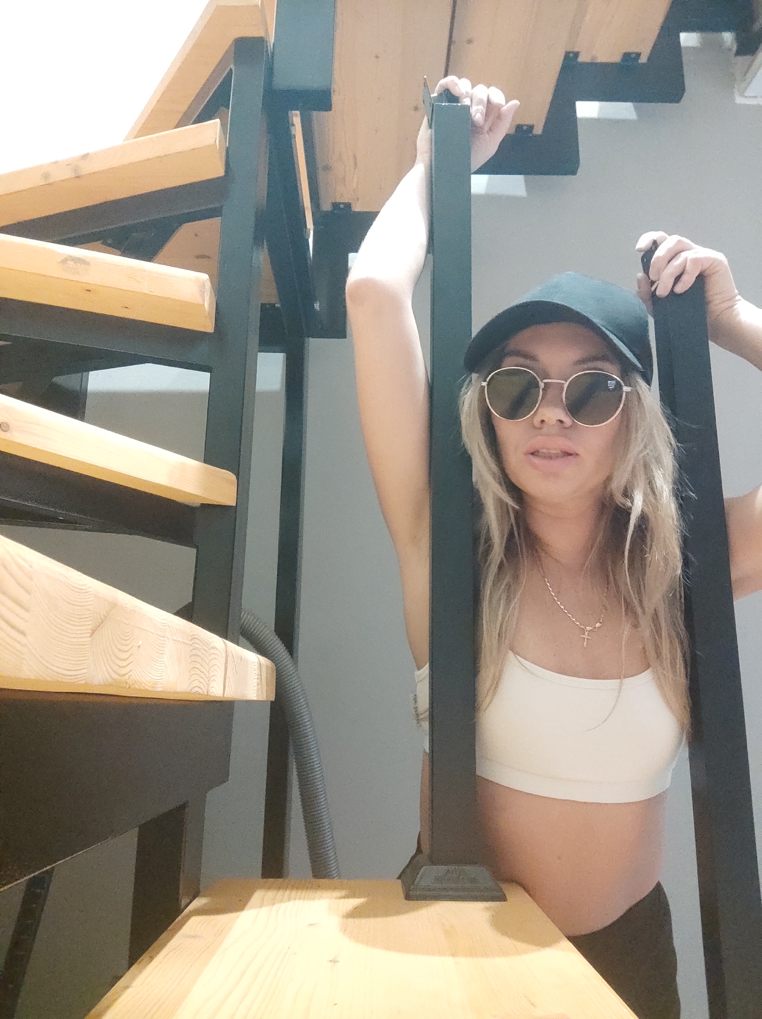 A woman wearing a white sports bra, dark pants, a black baseball cap, and round sunglasses stands next to a wooden and black metal staircase. The staircase is comprised of light-colored wood steps attached to a black metal frame. The woman holds onto the black metal uprights of the staircase with both hands. A gray hose is visible behind the staircase. The background wall is a neutral gray color. The location is inside a building, not specified within the city of Votkinsk, Russia.