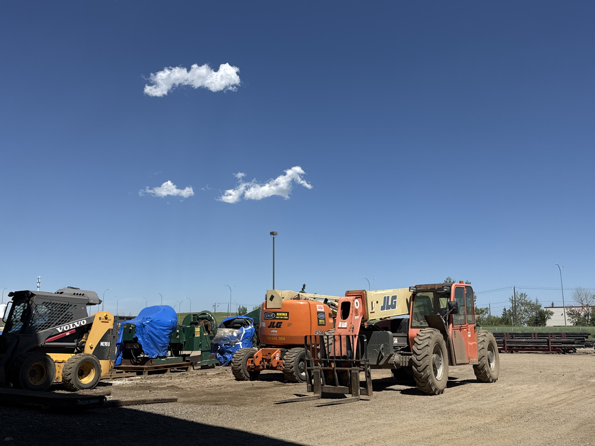 The image depicts an outdoor scene on a sunny day with a clear blue sky and scattered white clouds. The ground is covered in gravel and dirt. In the foreground, to the left, a yellow and black Volvo skid steer loader is parked. To the right of the skid steer, there are several pieces of industrial equipment, including a green machine covered partially by a blue tarp, and another large piece of machinery, possibly a generator, also covered in a blue tarp. Further to the right is a large orange JLG telehandler with a forklift attachment. In the background, there are streetlights, buildings, and stacks of what appear to be metal pipes or beams. The overall impression is that of a construction or industrial equipment storage yard. Based on the presence of the specific equipment and the general environment, this scene is likely located in an industrial area within the city of Calgary, Canada. No people are visible in the image.