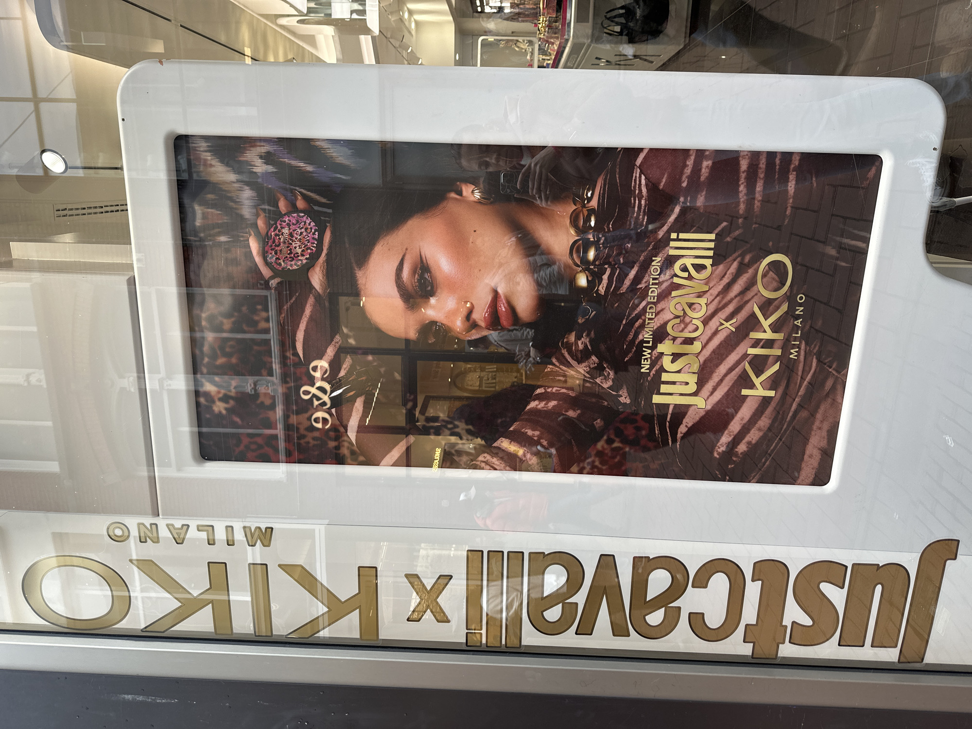 A storefront in Amsterdam, Netherlands, displays an advertisement for a collaboration between Just Cavalli and Kiko Milano. The ad features a model with dark hair and tanned skin wearing a patterned top and holding a makeup compact. The text "Just Cavalli x Kiko Milano" is prominently displayed in gold lettering. The ad is framed in a white rectangular display. The reflection on the glass includes elements of the street and the store's interior.