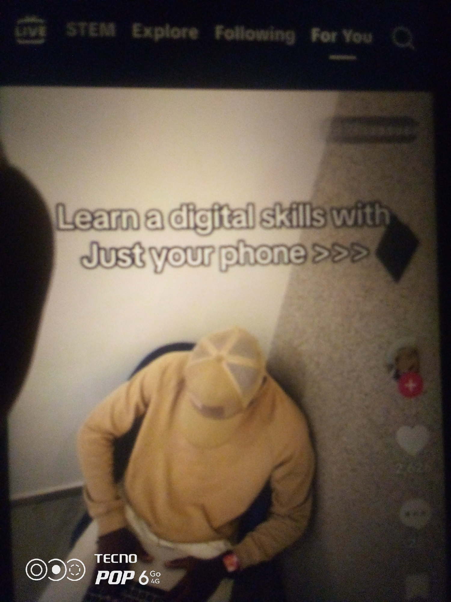 The image displays a screen, likely from a mobile device, featuring a social media interface. At the top, a navigation bar shows options like "LIVE," "STEM," "Explore," "Following," and "For You," alongside a search icon. A prominent text overlay reads "Learn a digital skills with Just your phone >>>". Below this, a person wearing a yellow hoodie and a matching baseball cap is seated, partially visible. The background is a textured wall with some icons on the right side, including a heart symbol with a number "2,626" below it, and a plus icon within a red circle. In the bottom left corner, a logo that reads "TECNO POP 6 Go 4G" is visible. The image is blurry and appears to be taken in low light conditions. The location in Howayi, Nigeria, is not determinable from the visual content of the image.