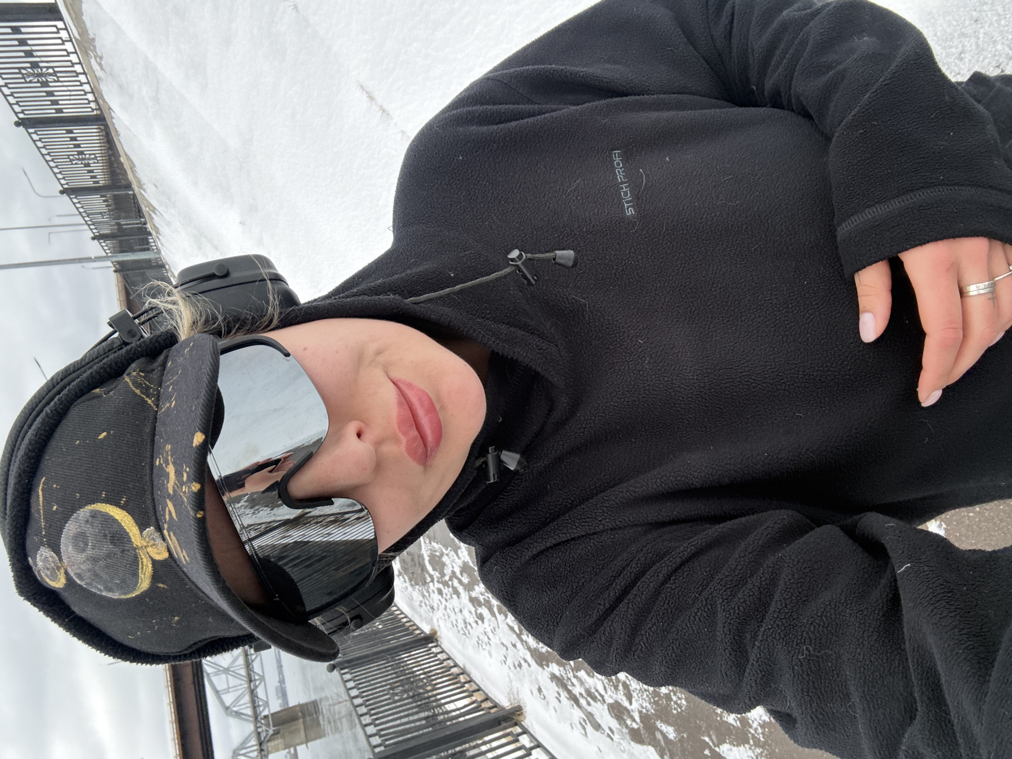 A woman in a black fleece jacket and decorated black hat with sunglasses stands on a bridge, likely the Vinogradovsky Bridge in Krasnoyarsk, Russia, given the bridge's design and the snowy surroundings, suggestive of the Siberian climate. The backdrop consists of snow-covered ground and the distinctive metal railings of the bridge. The woman is facing the camera, suggesting a self-portrait or a photo taken by someone nearby.