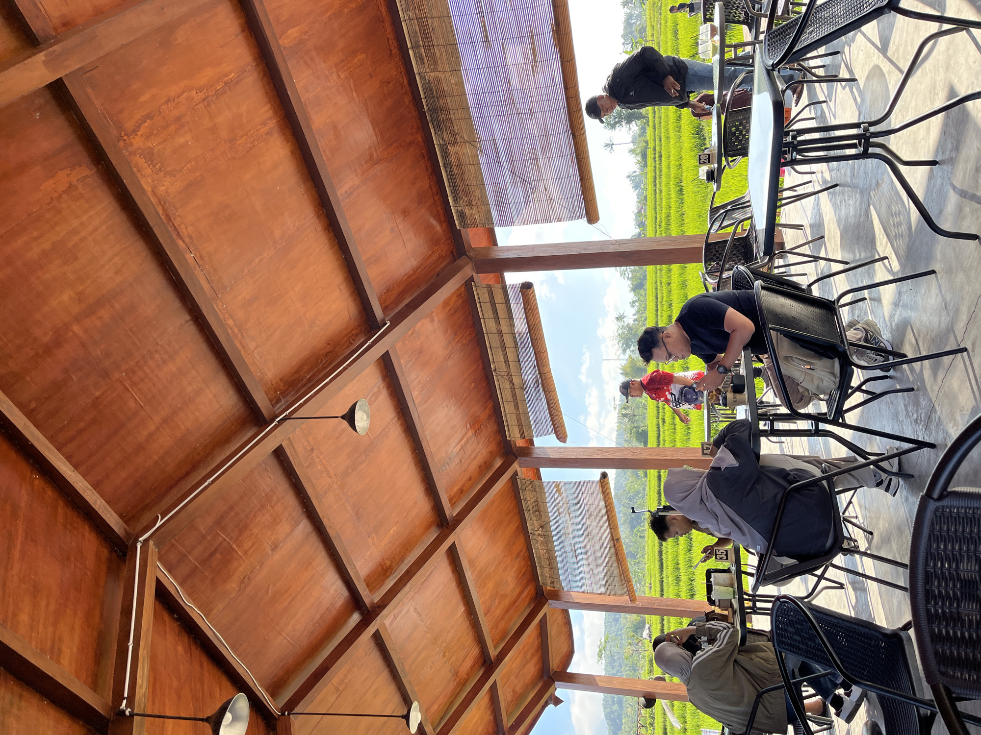 The image depicts an outdoor seating area at a cafe, likely located in Oro-oro Ombo, Indonesia, with a view of lush green rice fields under a bright, sunny sky. The structure is an open-air pavilion with a warm wooden roof supported by pillars, featuring woven bamboo blinds on the sides. Several tables and chairs, made of metal and with woven seats and backs, are arranged on a concrete floor.

In the foreground, several individuals are seated at tables, engaged in conversations and using their phones. A woman in a hijab and a green jacket is seated at a table on the left, looking at her phone. Next to her, a woman in a grey hijab is also seated at a table. In the center, a man wearing glasses and a black t-shirt is looking down at his phone. Further back, a man in a red shirt stands near a table. To the right, a man in a black jacket and jeans is standing, looking towards the rice fields.

The environment outside the pavilion consists of vibrant green rice paddies, suggesting a rural or semi-rural setting. The sky is a clear blue with some scattered clouds. The overall atmosphere appears relaxed and casual, with patrons enjoying their time at the cafe amidst the natural scenery.