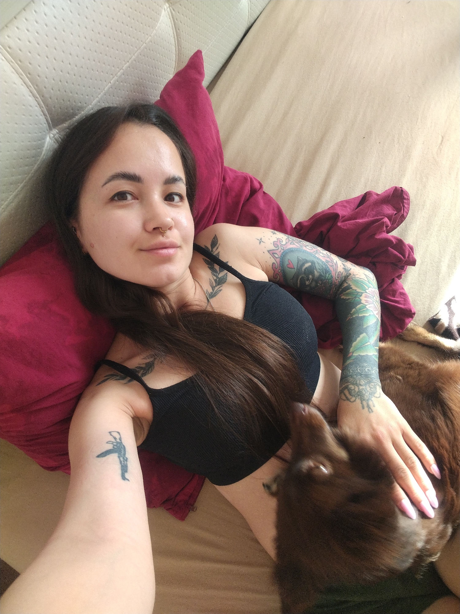 A young woman with long dark hair and a nose ring is lying on a bed with a dog. She is wearing a black tank top and has several tattoos on her arms. The dog is brown and appears to be resting on her. The scene is indoors, likely a bedroom, with a textured headboard visible in the background. A dark red pillow is positioned behind the woman's head. The lighting suggests it is daytime. The woman is looking at the camera with a relaxed expression, and her hand is resting on the dog. The overall mood is casual and intimate. No visible text is present in the image.