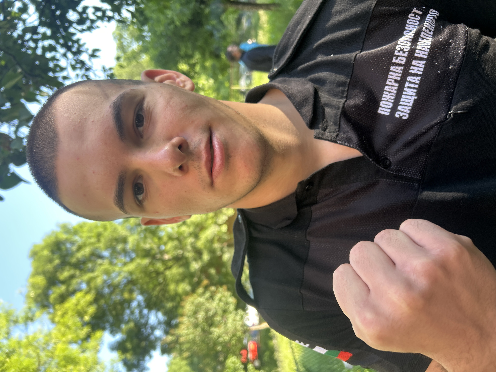 A close-up, slightly tilted photograph shows a young adult male outdoors. He has a shaved head, light complexion, and neutral expression. He is wearing a black uniform shirt with white Cyrillic text on the upper right chest. A patch with the Bulgarian flag is visible on the lower portion of the uniform. His right hand is clenched into a fist. The background includes trees and a blue sky. Another individual is partially visible in the background. The location appears to be a park or green space.