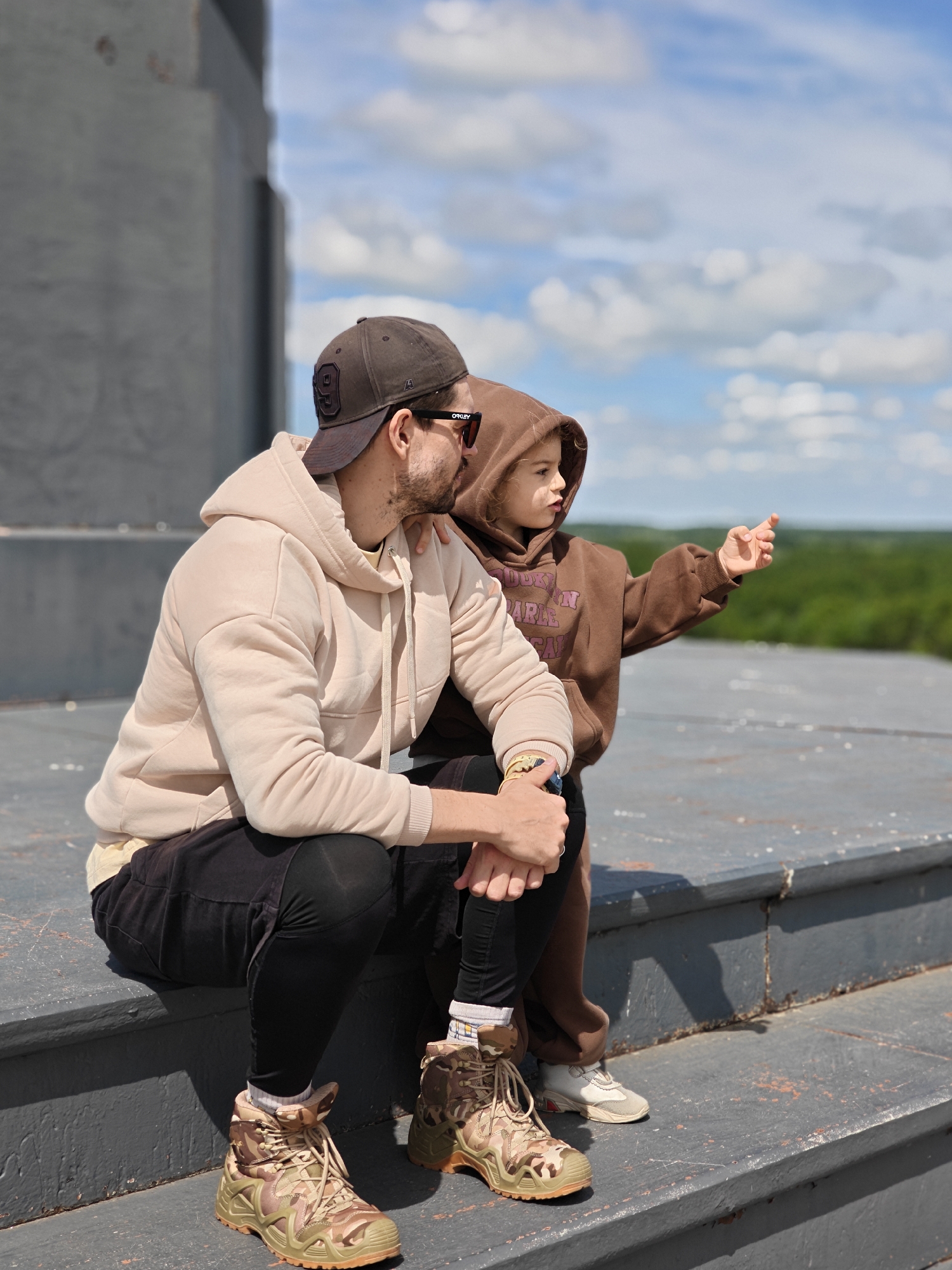 The photograph shows a man and a child, likely a father and daughter, sitting on a gray, multi-tiered concrete structure against a backdrop of a blue, partly cloudy sky and a green treeline. The structure's tiered design suggests it may be part of a memorial or monument.
The man is wearing a light beige hooded sweatshirt, black pants, and camouflage pattern boots. He has a baseball cap on backward and is wearing sunglasses. He is sitting with his legs bent, arms resting on his knees. The child is wearing a brown hooded sweatshirt and is standing next to the man, pointing her right arm out toward the horizon.
The location of the photo is Borodino, Russia. The details include the concrete structure, the clothing and footwear worn by the subjects, the relative positions of the two people, and elements of the surrounding environment like the sky and trees. There is a clear focus on the two individuals in the foreground.