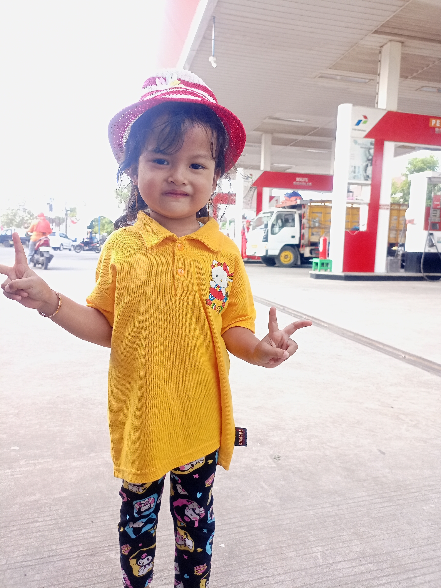 A young girl stands at a gas station, smiling at the camera. She is wearing a yellow polo shirt with a Hello Kitty graphic on it and a red and white striped hat. Her pants are black with a cartoon print. She holds up two peace signs, one with each hand. In the background, gas pumps and a truck are visible, along with other vehicles and buildings, suggesting the location is a busy area. The overall setting indicates a typical day at a gas station, possibly in a public or commercial district of Juwana, Indonesia.