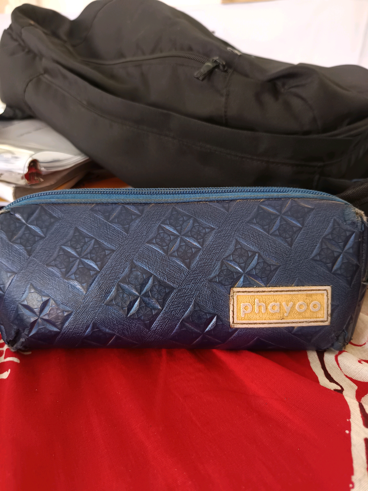 A blue pencil case with a textured pattern and a yellow label reading "phayoo" is placed on a red fabric surface. Behind the pencil case, a black backpack is visible. The scene appears to be indoors. There is no discernible location in the city of Labuhan, Indonesia.