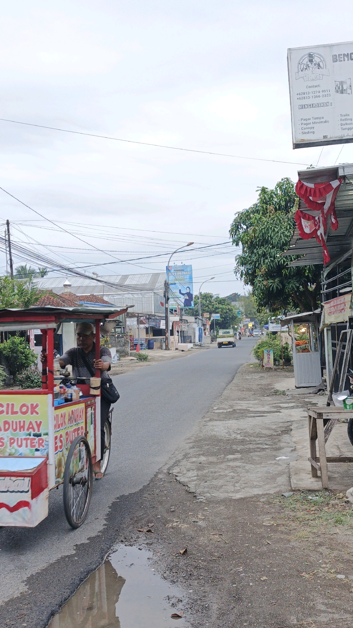 In the city of Parigi, Indonesia, a street scene unfolds under an overcast sky. A man, wearing a cap and a patterned shirt, stands beside a colorful food cart. The cart, adorned with red and white banners and text reading "CILOK ADUHAY ES PUTAR", is parked on the asphalt road. The cart has two wheels and is equipped with various containers and what appears to be a small stove. To the right of the road, a sidewalk made of concrete slabs is visible, with some debris and vegetation. Beyond the sidewalk, several small businesses and shops line the street. Overhead, a network of power lines crisscrosses the sky. In the background, a light-colored truck drives away from the camera, and other vehicles and buildings suggest a typical urban environment. A large billboard with text and an image of a woman is also present further down the road. The overall scene depicts a busy street in a commercial area of Parigi.