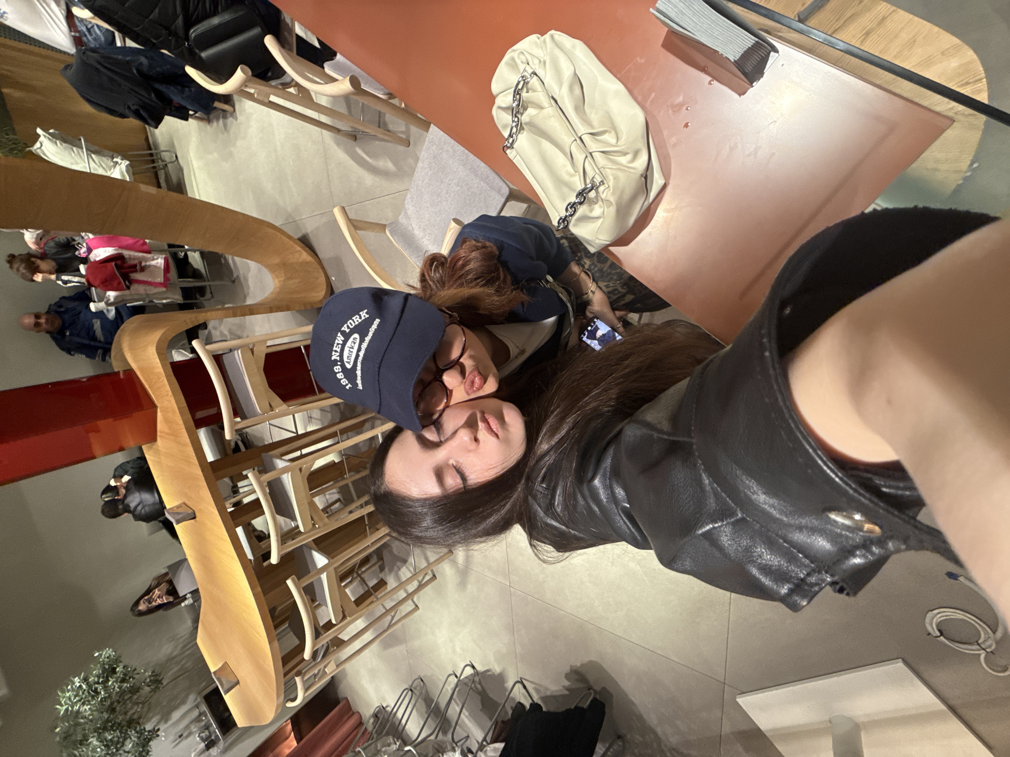 The image shows a tilted, indoor shot of two women posing for a selfie. One woman is wearing a black leather jacket and has long, dark hair. The other is wearing a blue New York baseball cap and glasses. They are sitting at a table. On the table is a cream-colored purse with chain detailing and a stack of papers.
The background consists of a light-colored tile floor, wooden chairs, and a wooden shelving unit. Some items are on the shelving unit. A small tree in a pot can be seen in the distance.
Based on the architectural style and decor, this is likely a commercial space in Yerevan, Armenia.