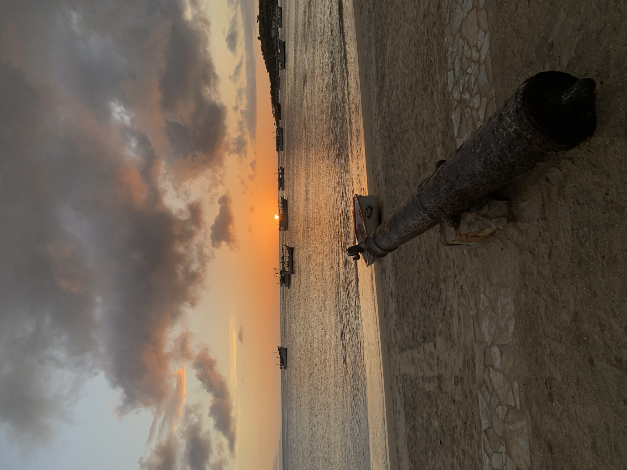 A sunset over the ocean in Juan Griego, Venezuela, casts a warm orange glow across the sky, illuminating scattered clouds. The calm water reflects the colors of the sunset. Several boats are anchored in the bay, some closer to shore and others further out. On the sandy beach in the foreground, a weathered cannon rests on a stone base. A small white boat with an outboard motor is pulled up onto the sand near the water's edge. The scene appears tranquil, with no people visible. The overall impression is one of peaceful natural beauty, with a historical artifact adding a touch of character to the landscape.
