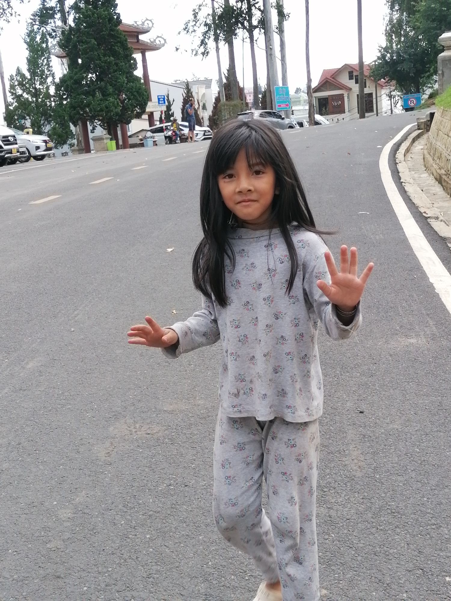 A young Vietnamese girl with long black hair stands on a paved road in Da Lat, Vietnam, facing the camera and raising her right hand with fingers spread. She is wearing a gray long-sleeved shirt and matching pants, both adorned with a pattern of small pink and blue flowers. Her left hand is slightly extended to the side. Behind her, the road curves to the right, bordered by a stone retaining wall and a white stripe. In the background, several tall trees with dark green foliage are visible, along with buildings and parked cars. The sky appears overcast. The girl is the central subject, positioned in the foreground of the image.