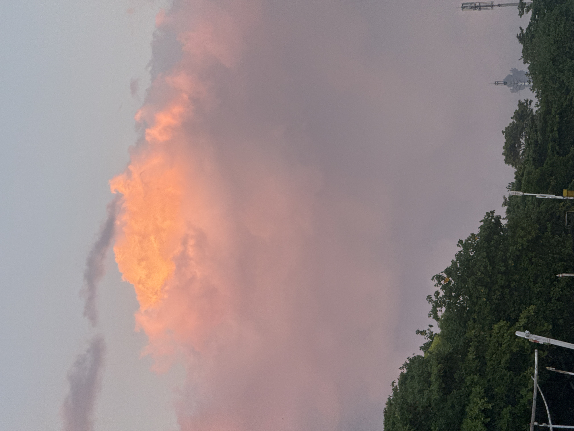 This is an eye-level, medium shot photograph taken during the golden hour, showcasing a dramatic sky filled with vibrant pink and orange clouds. The clouds dominate the upper portion of the image, with a particularly bright, illuminated mass of pink and orange cloud positioned centrally. Lower down, the sky transitions to a soft, hazy pink and then to a pale, muted blue.
In the foreground, a dense canopy of dark green trees runs horizontally across the bottom of the frame. The foliage appears lush and full. Peeking out from behind the trees on the right side of the image are two modern telecommunication towers, one taller and more prominent than the other. In the distance, silhouetted against the hazy sky, is a distinctive, multi-tiered tower structure, possibly a monument or a unique building, giving a hint of urban landscape. There are no people or animals visible in the scene. The overall atmosphere is serene and picturesque, with the vibrant clouds creating a striking contrast against the natural and man-made elements of the landscape. Given the presence of telecommunication towers and the distinctive architectural silhouette, this scene is likely located in a developed area near Kuta, Indonesia, possibly on the outskirts or in a less densely populated part of the city, where the natural elements of trees and sky are still prominent.