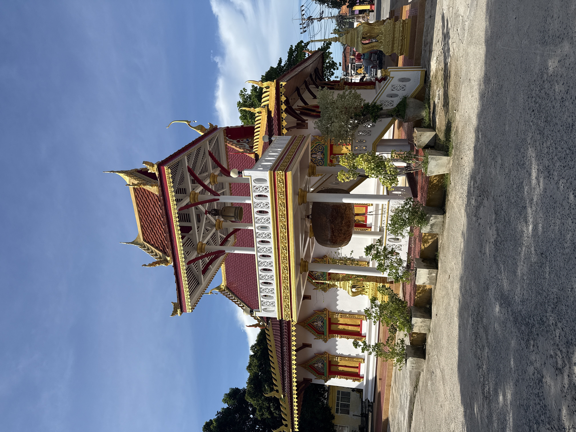 The image shows a Buddhist temple with ornate golden trim and a red tiled roof, located in Baan Lamai, Thailand. The temple is painted white and features decorative windows with red and gold accents. In front of the temple, there is a large bell hanging from a covered structure. Several potted plants are arranged in front of the temple. The sky is blue with scattered clouds. The ground in the foreground is paved with asphalt, and shadows from the temple and plants are visible. A street with a few vehicles and utility poles is visible in the background, suggesting an urban or semi-urban setting. There are no people visible in the image.