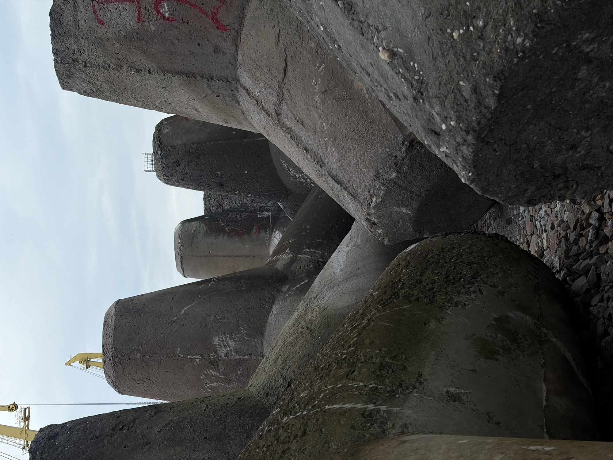 The image depicts a close-up, upward-angled view of several concrete tetrapods. These tetrapods are grey in color and appear to be weathered. The scene includes the presence of a crane in the background. The tetrapods are located in Burgas, Bulgaria. The tetrapods are partially resting on or surrounded by stones. Graffiti in red is visible on one of the tetrapods.