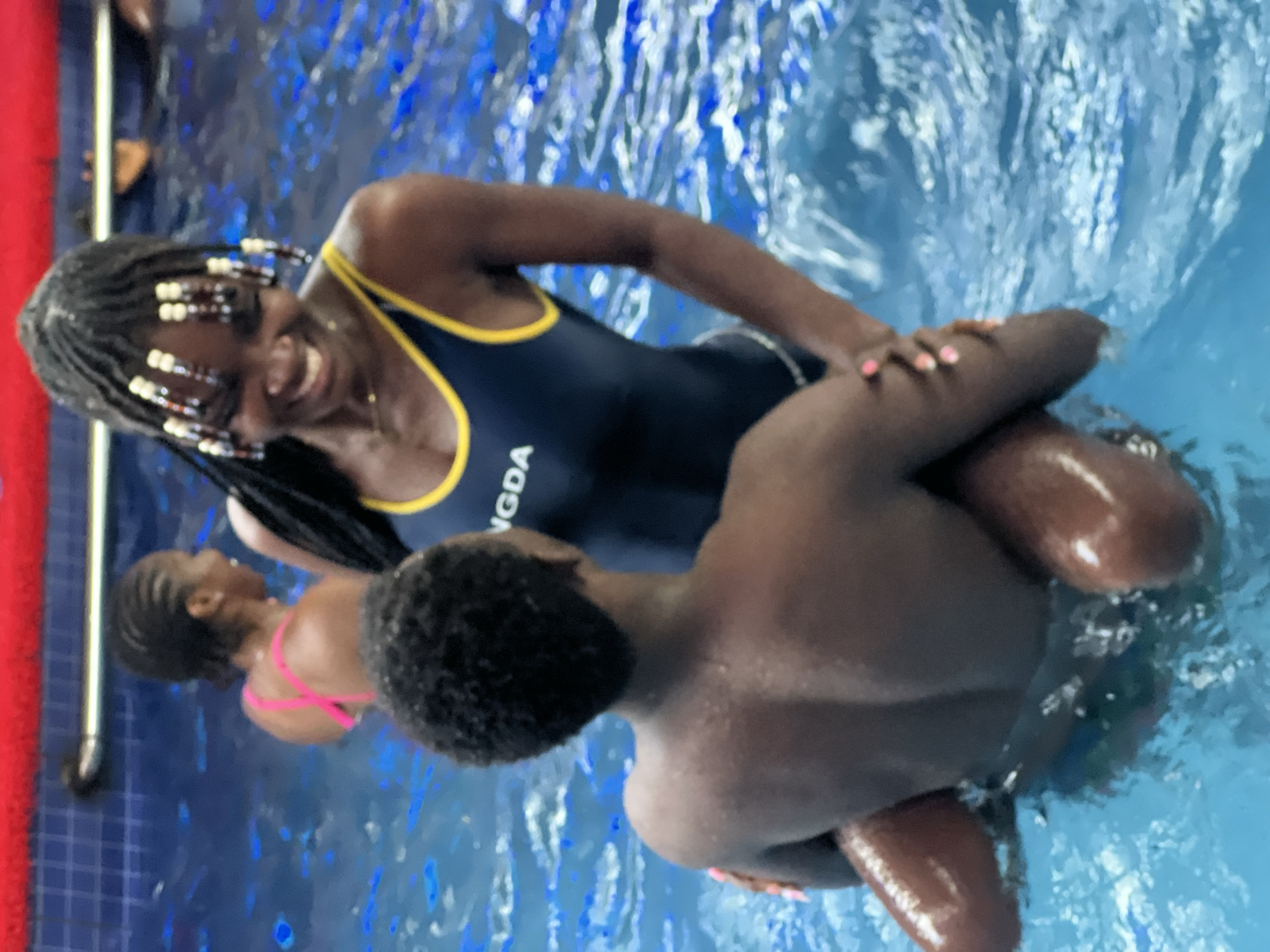 The image shows a young Black couple in a swimming pool. The woman is wearing a navy blue swimsuit with yellow trim and has her hair braided with beads. She is smiling and looking at the man. The man is holding her in his arms, and they are both in the water. The swimming pool appears to be indoors, with blue tiled walls and a red coping around the edge. The water is clear and blue, with ripples on the surface. The overall impression is one of playful affection between the couple. The location is a swimming pool in Obosi, Nigeria.