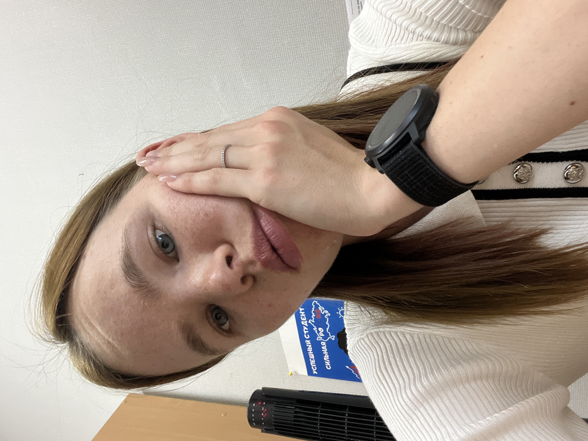 A close-up, angled view presents a person with fair skin and light brown hair. They are wearing a white, ribbed top with black stripes and silver buttons visible near the right shoulder. A black smartwatch is on their left wrist. The left hand, adorned with a silver ring on the ring finger, is near their face, partially obscuring it.
In the background, a desk is visible along with a wall. A poster, seemingly in Russian, is partially visible, bearing the phrase "УСПЕШНЫЙ СТУДЕНТ" (Successful Student) and the word "РОССИЯ" (Russia). A black, electric heater is also visible on the desk.
The location is potentially Chaikovsky, Russia, given the Cyrillic script and context. The scene suggests a personal space, possibly a student's room or study area.