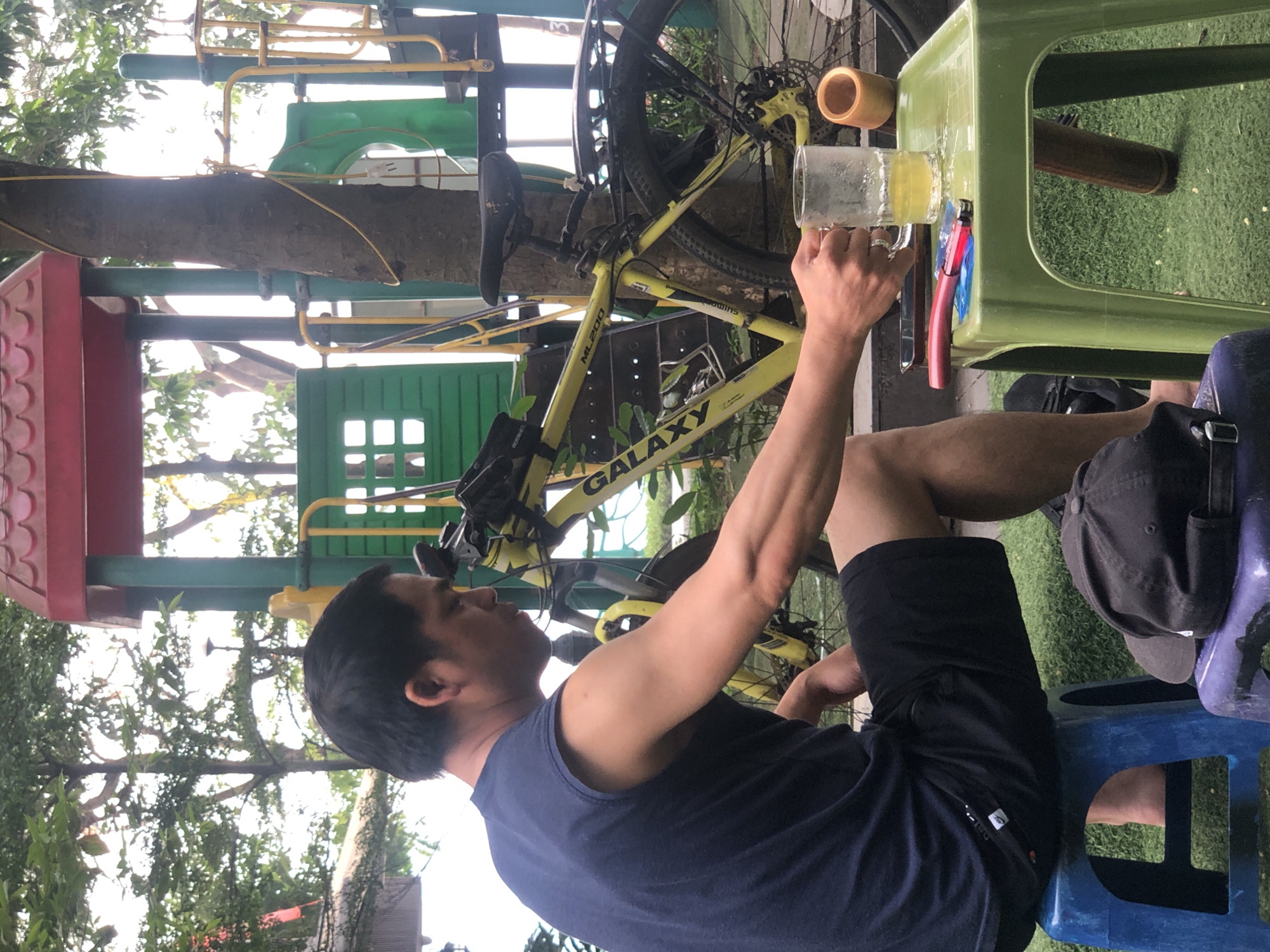 A medium shot captures a man holding a glass of beer at an outdoor cafe in Hà Nội, Vietnam. He is dressed in a navy tank top and black shorts, seated on a blue plastic chair. A black baseball cap rests beside him on the chair. His face is in profile, looking towards the glass of beer in his hand. A yellow bicycle leans against a nearby structure. The table he is sitting at is light green and holds a cylindrical object beside the glass. In the background is a playground structure with red, green, and yellow elements, partially obscured by the foliage of trees. The ground is covered in artificial grass.