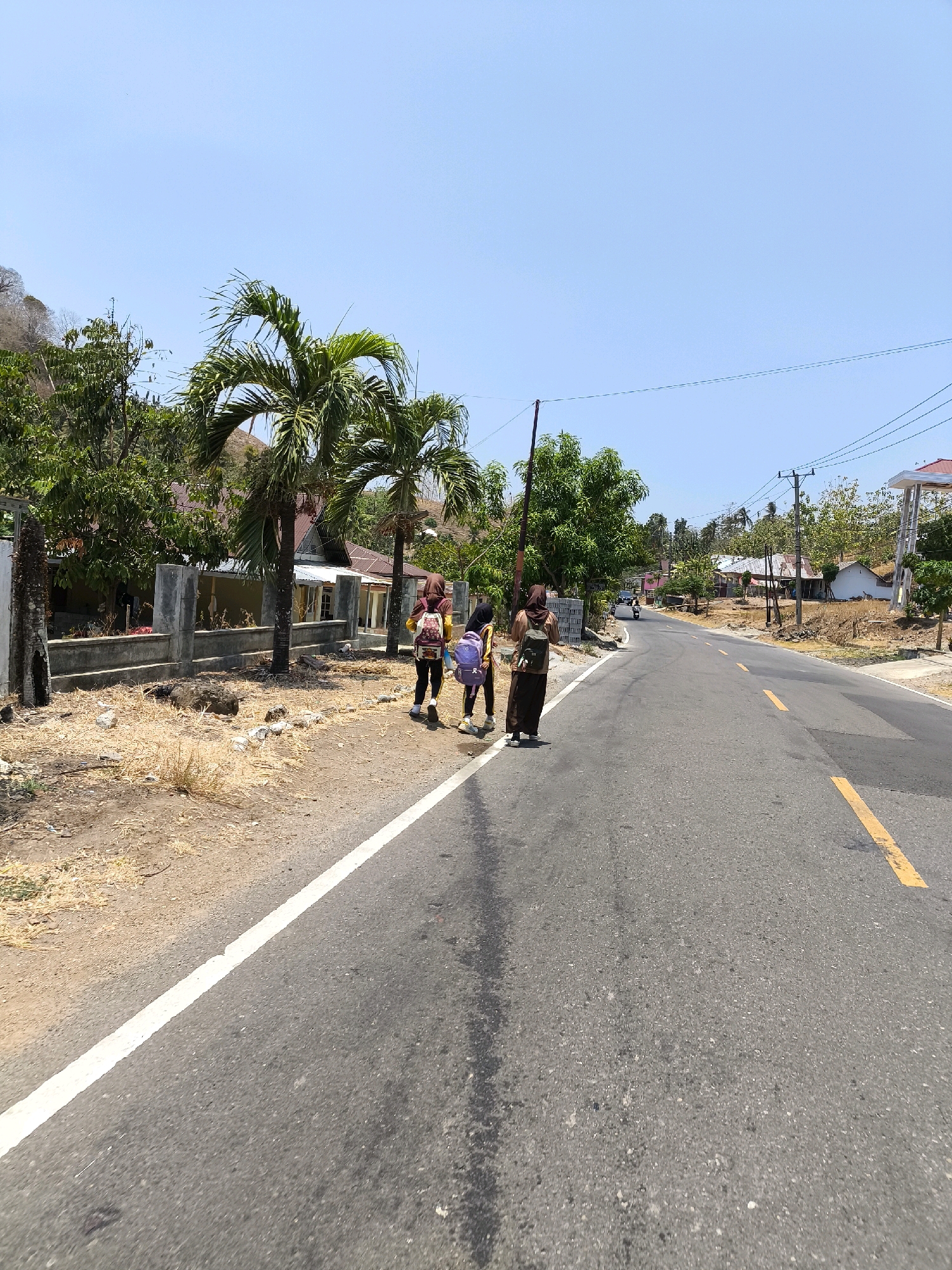 The image depicts a paved road in Kwandang, Indonesia, on a clear, sunny day. The road has a white line marking the shoulder and a yellow line down the center. On the left side of the road, there is dry, yellowish grass and a concrete fence. Behind the fence, there are palm trees and houses with red roofs. Three individuals are walking along the side of the road. The person furthest to the right is wearing a brown hijab and a long brown skirt, carrying a backpack. To their left, a person in a yellow shirt and dark pants with a backpack walks ahead, followed by another person in a yellow shirt and dark pants also wearing a backpack. In the distance, there are more houses, trees, and utility poles with wires. A motorcycle is visible on the road further down. The overall environment suggests a rural or semi-rural setting in a tropical climate.