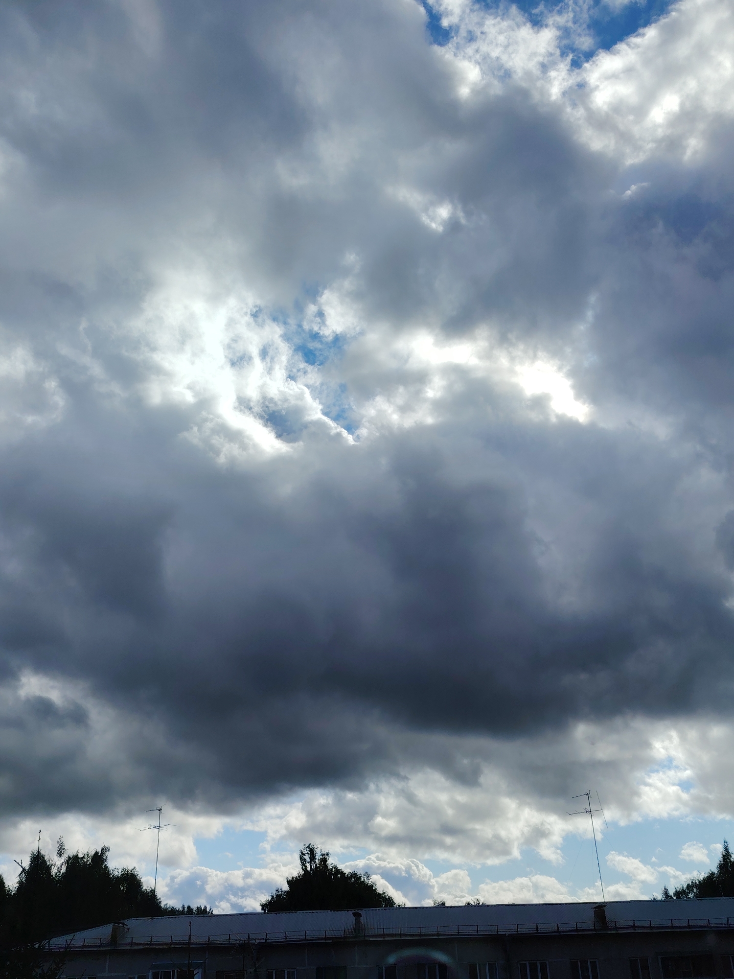 A wide shot captures a building's roofline in Konakovo, Russia, against a dramatic sky filled with dark, heavy clouds. A few breaks in the clouds reveal patches of blue sky. The roof appears metallic and has several antennas attached to it. Silhouetted trees line the horizon, obscuring the ground level. The scene is overcast and suggests an impending storm.