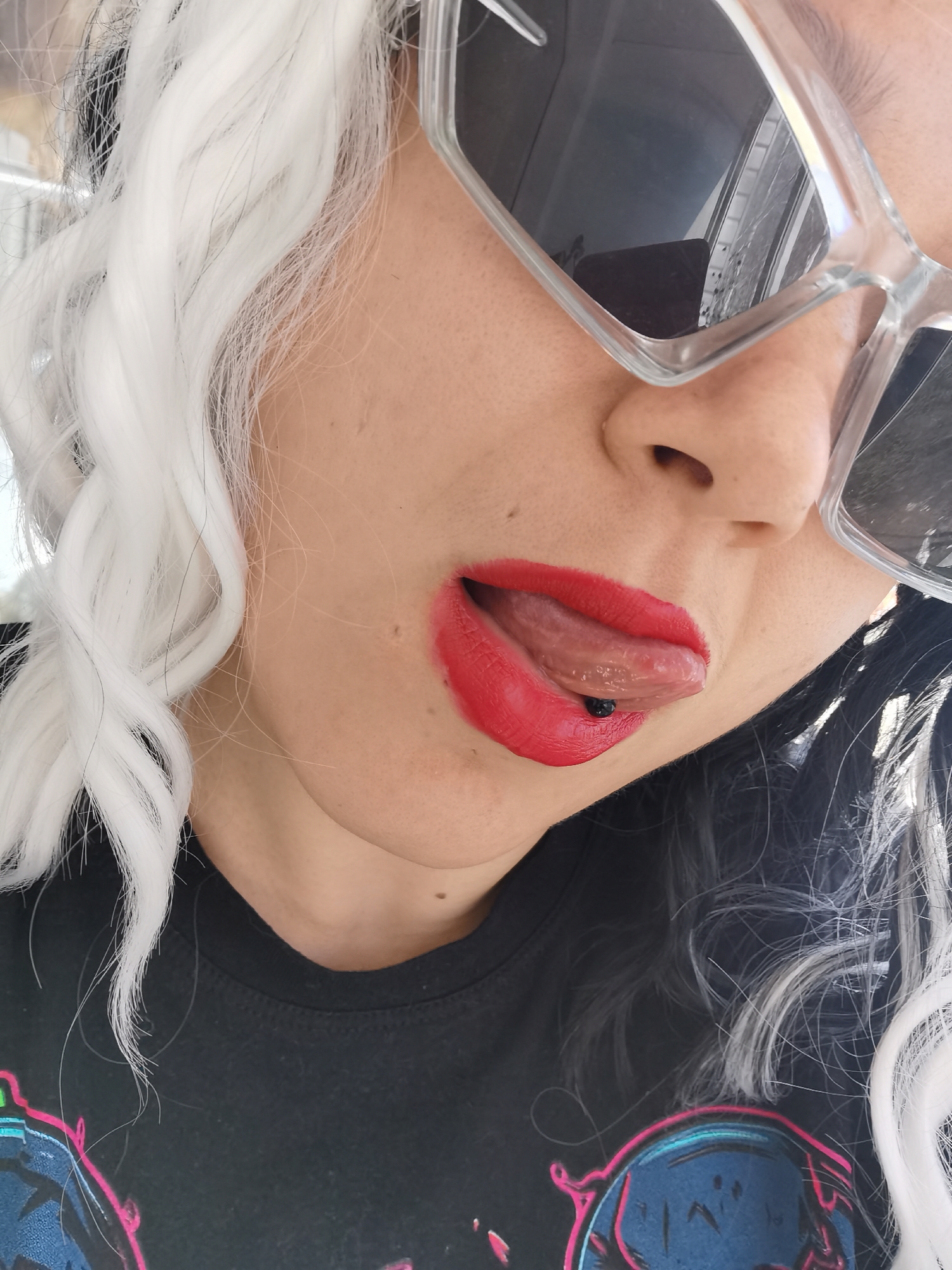 A close-up, mid-shot portrait features a person with fair skin, captured from the nose down and from the upper chest. The person is wearing large, clear-framed, rectangular sunglasses with dark lenses, which obscure the upper right side of their face. Below the sunglasses, a nose is visible, and the mouth is open with the tongue playfully sticking out. The lips are adorned with bright red lipstick, and a small, black, spherical piercing is visible on the underside of the tongue.
The person has striking two-toned hair, appearing to be white and black. Wavy, white hair cascades down the left side of the frame, contrasting with darker hair visible on the right and behind the white strands. The person is wearing a black T-shirt with a colorful, partially visible graphic design on the chest, featuring shades of pink and blue. The overall mood conveyed is playful and expressive. The lighting is bright, suggesting daytime conditions. The location context provided is Chaikovsky, Russia, though no specific visual cues confirm this within the image. There is no visible text.