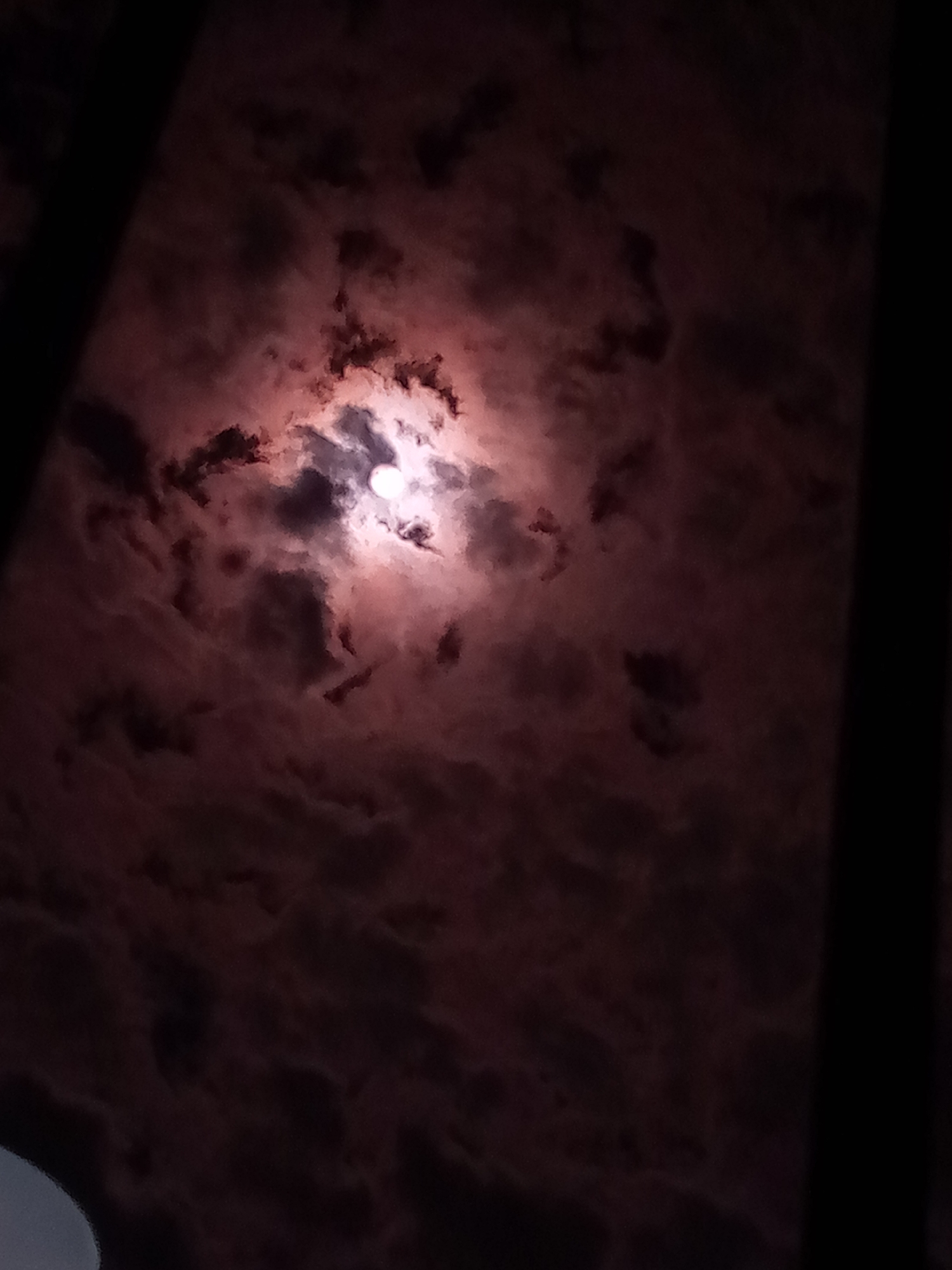 The image presents a dramatic nocturnal scene, focusing on a bright, partially obscured moon amidst a heavily clouded sky. The moon, a luminous white sphere, is positioned towards the upper-center, radiating light that intensely illuminates the surrounding clouds with a striking reddish-pinkish hue. These vibrant, warm tones contrast sharply with the deep black of the sky and the darker, unlit clouds that appear as shadowy forms scattered across the frame.
The time of day is clearly night, characterized by the dark backdrop and the moon being the primary light source. The weather conditions are cloudy. No people are visible in the image.
Dark, vertical bars flank the left and right edges of the frame, suggesting the view might be through a window or a similar architectural structure. A faint, light grey, curved shape is also visible in the bottom-left corner, further implying an indoor perspective looking out. There are no specific location cues beyond these hints. No text is visible in the media file. The overall impression is one of mystery and awe, stemming from the moon's ethereal glow through the cloud cover.