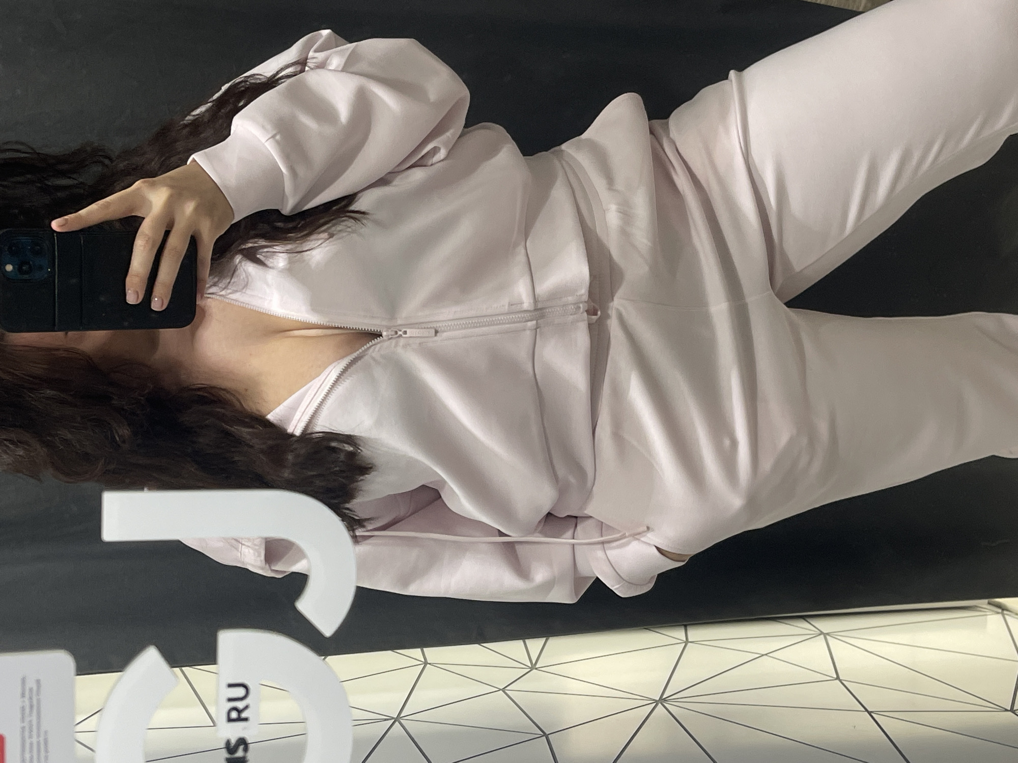 A person wearing a light pink zip-up tracksuit is taking a selfie in what appears to be a dressing room or clothing store. The person is holding a black smartphone in their left hand, partially obscuring their face. The person has dark hair. The background includes a gray or black wall and a floor with a white geometric pattern. In the lower left corner, part of a white sign or display with the letters "RUS" is visible. The location is likely a store somewhere in Moscow, Russia.