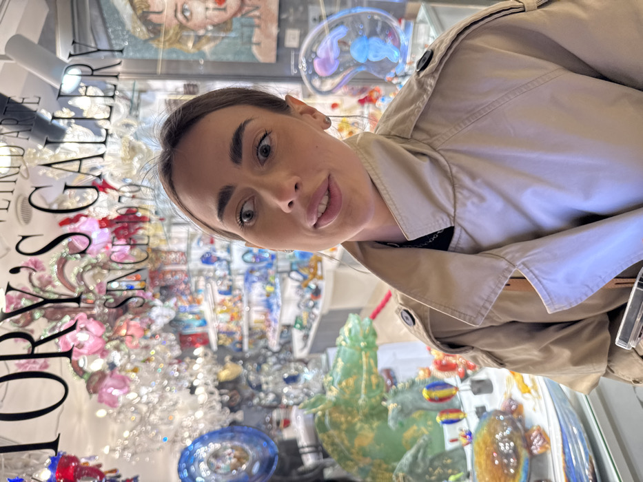 The image portrays a young adult female in the foreground, positioned outside a glass art gallery in Venice, Italy. She has light skin, dark eyebrows, and light brown hair, with wide eyes directed upwards and to the right, and her lips slightly parted. She is wearing a beige trench coat over a dark top, revealing a dark chain necklace. Her right hand, partially visible at the bottom of the frame, holds a smartphone. The background, visible through and reflected on the shop window, displays the interior of a brightly lit establishment identified by "VITTORY'S GALLERY, VETRO D'ARTE SINCE 1970" written on the glass. The gallery showcases numerous Murano glass items, including elaborate chandeliers with colorful floral accents, a large green and gold glass horse head sculpture, smaller colorful glass animals, abstract glass forms (one with a blue internal structure resembling a jellyfish), and a pop-art style painting of a blonde woman reflected on the right side of the window.