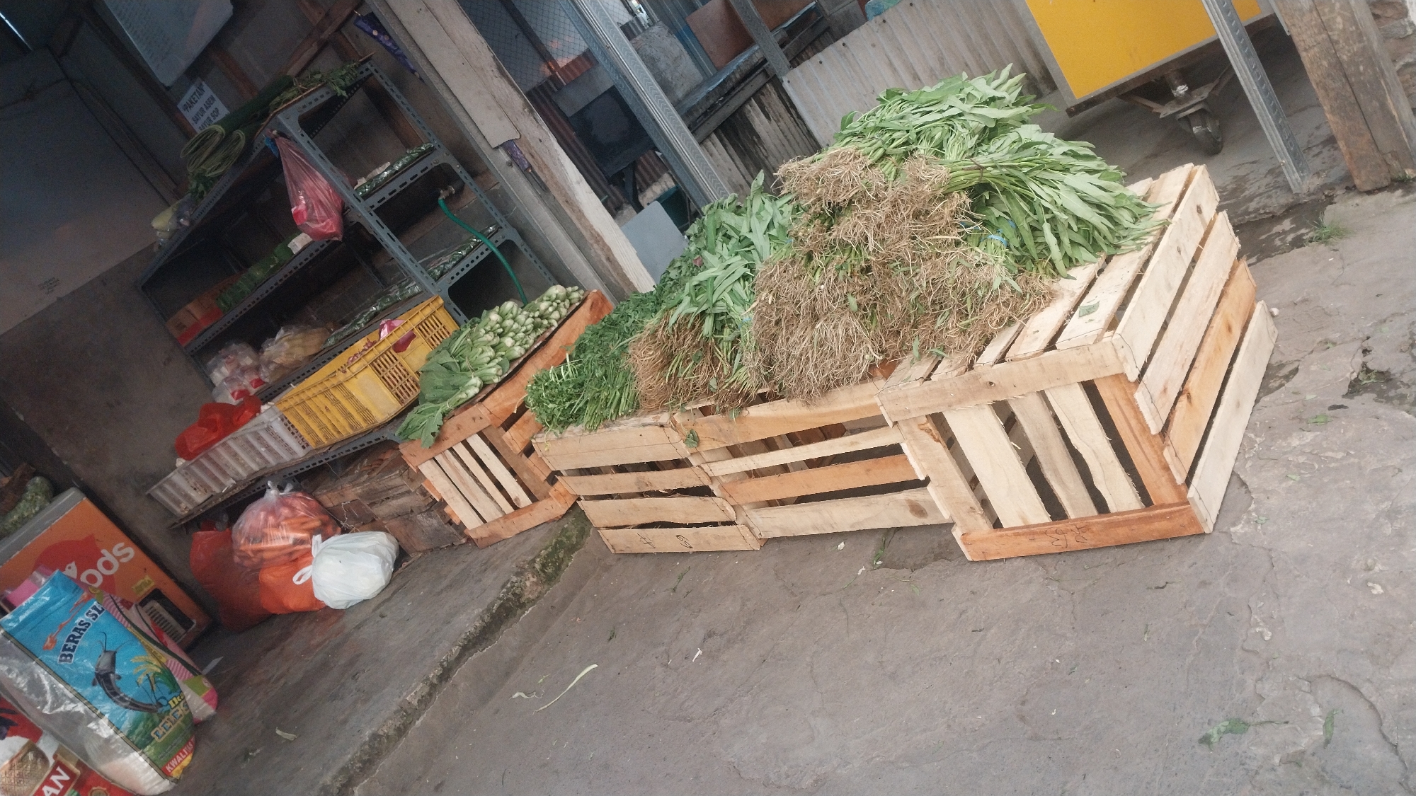 An outdoor market stall in Keboananom, Indonesia, features a display of fresh produce. In the foreground, several wooden crates are stacked, some filled with green leafy vegetables and bundles of roots. Behind the crates, a metal shelving unit holds more vegetables and some packaged goods. To the left of the crates, a bag of rice and some red bags containing vegetables are visible on the ground. The scene is viewed from a slightly elevated angle, with a concrete floor in the foreground.