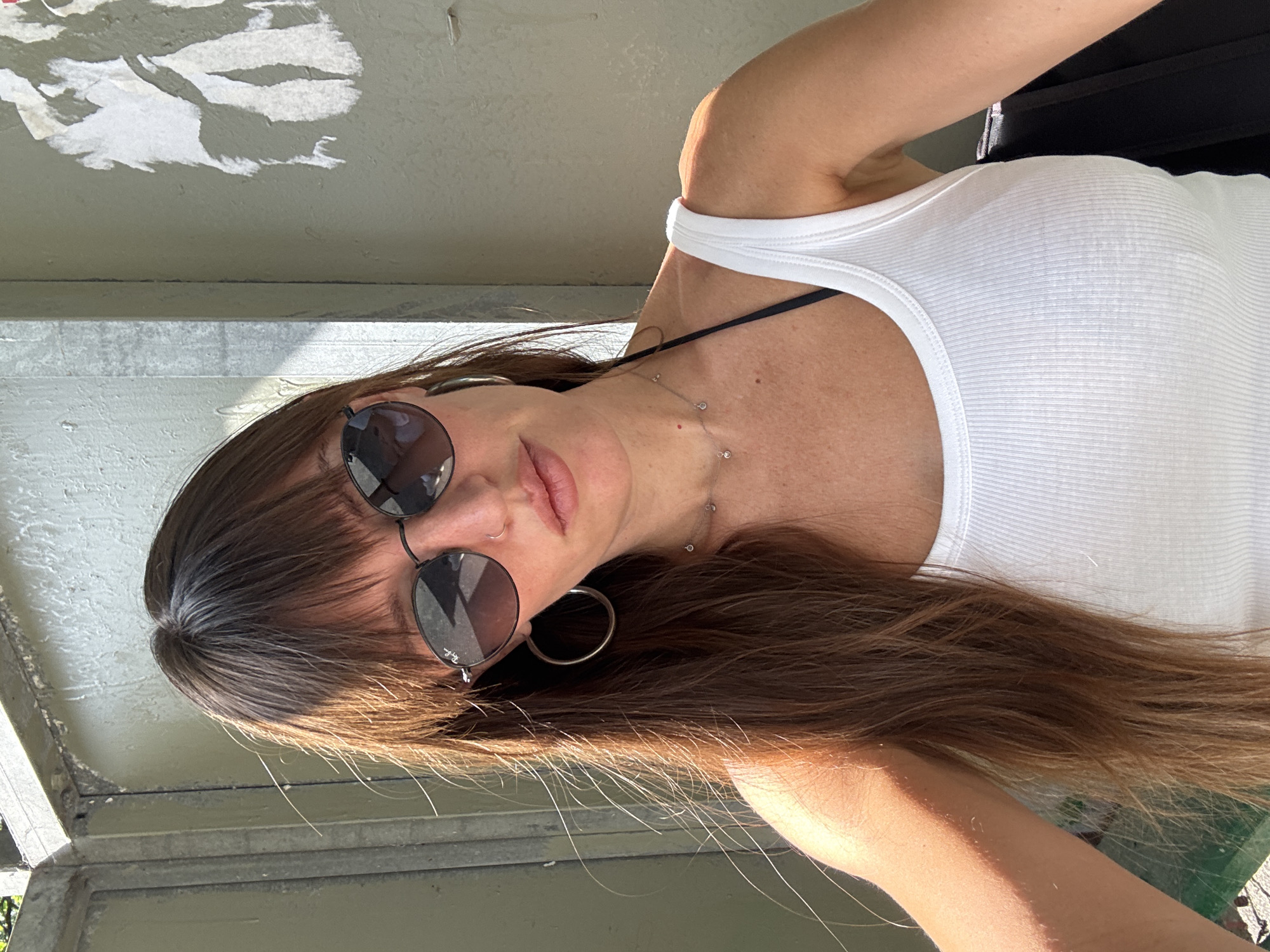 A woman with long brown hair and bangs, wearing sunglasses and a white tank top, poses for a selfie outdoors. She is wearing hoop earrings and a delicate necklace. The background appears to be a textured wall with torn paper adhered to it, and metal bars to the left. The lighting suggests it is daytime, with shadows cast on the wall. The location is not specified in the image data or the provided information.