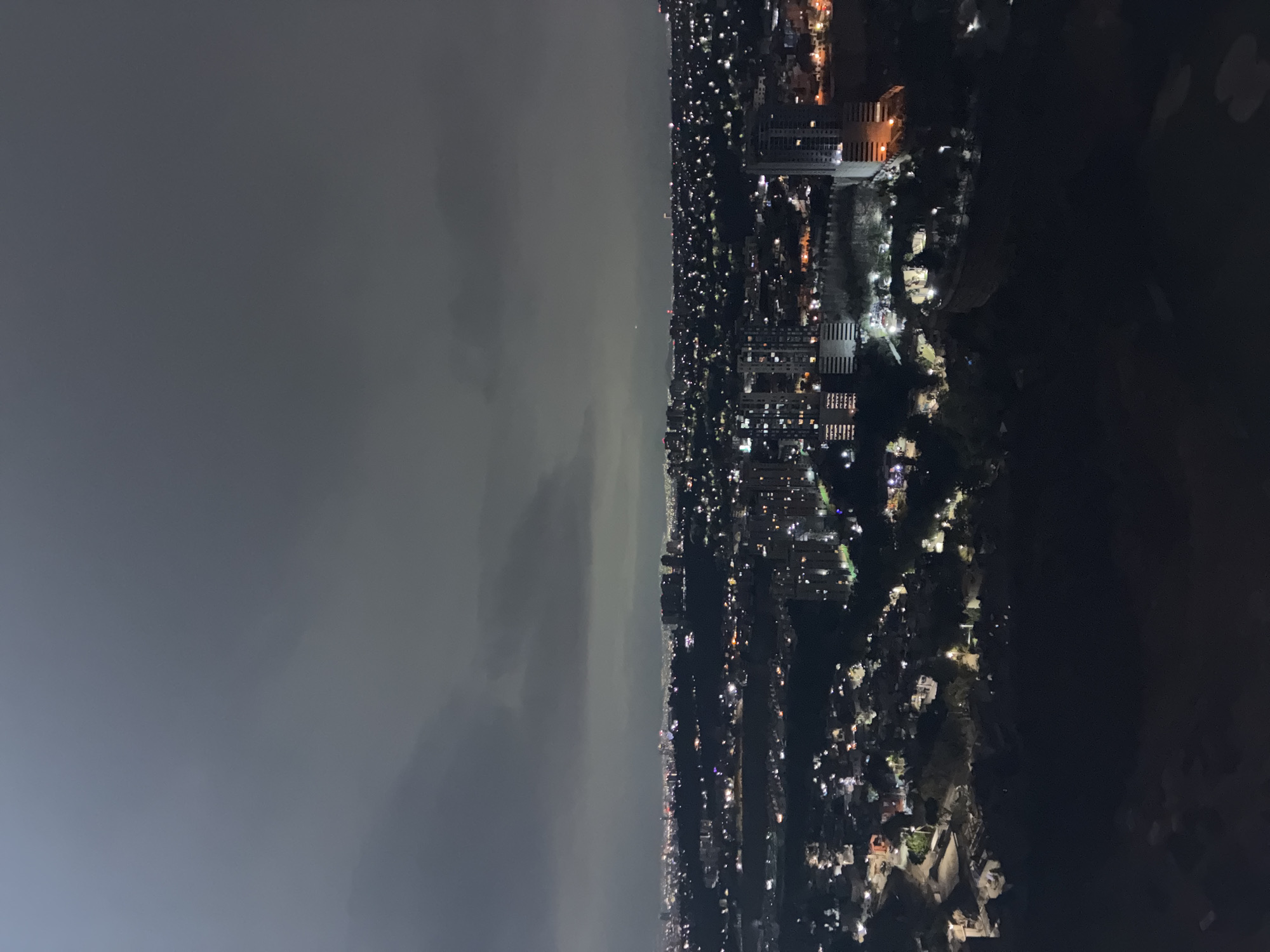 A high-angle, nighttime view of the city of Huixquilucan de Degollado, Mexico. The left portion of the image shows a cloudy or smoggy sky. The right portion presents the city, illuminated by streetlights and building lights. Buildings of varying heights and sizes are visible. The lower right corner is consumed by darkness, possibly indicating a less populated area.