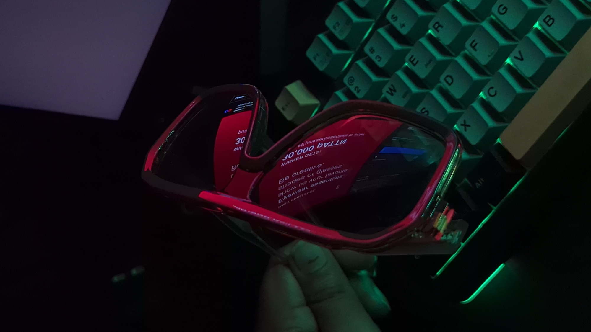 A close-up shot shows a pair of red-framed glasses held by a person. The glasses reflect a screen displaying red text, including the word "30,000." To the right of the glasses is a mechanical keyboard with backlit green keys. The background is dark, and the lighting is predominantly green and red. The image does not depict a specific location in Dhaka, Bangladesh.