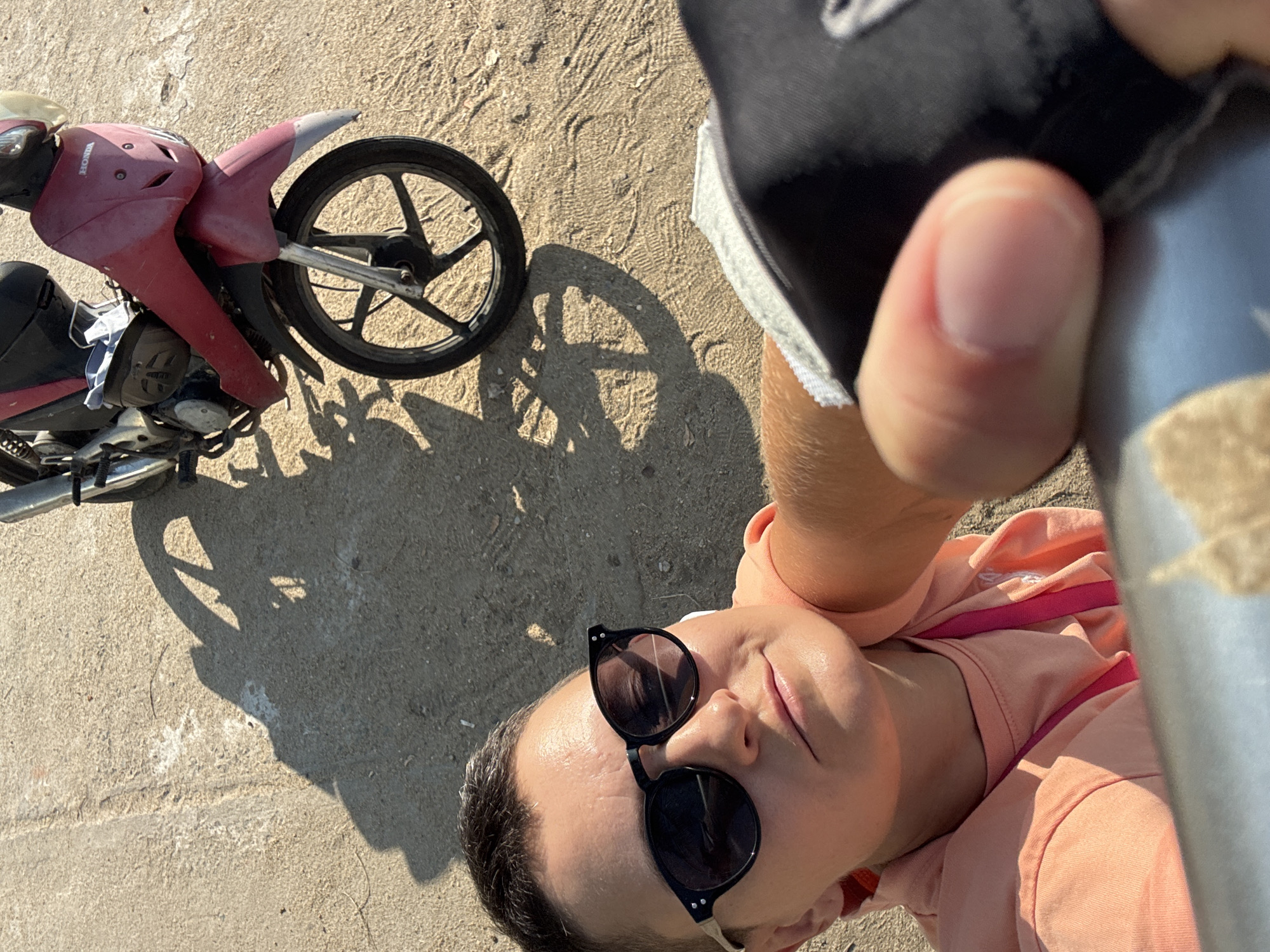 An overhead shot captures a person taking a selfie with a red Honda motorcycle parked on a concrete surface. The person, wearing a peach-colored hoodie and black sunglasses, is in the foreground, with their arm extended to hold the camera. Their hand, covered by a black glove with white trim, is visible near the bottom right corner. The motorcycle's front wheel and part of its body are in the mid-ground, casting a shadow on the ground. The background consists of the textured concrete, possibly a street or sidewalk, in Nha Trang Ward, Vietnam, bathed in the bright sunlight of a clear day.