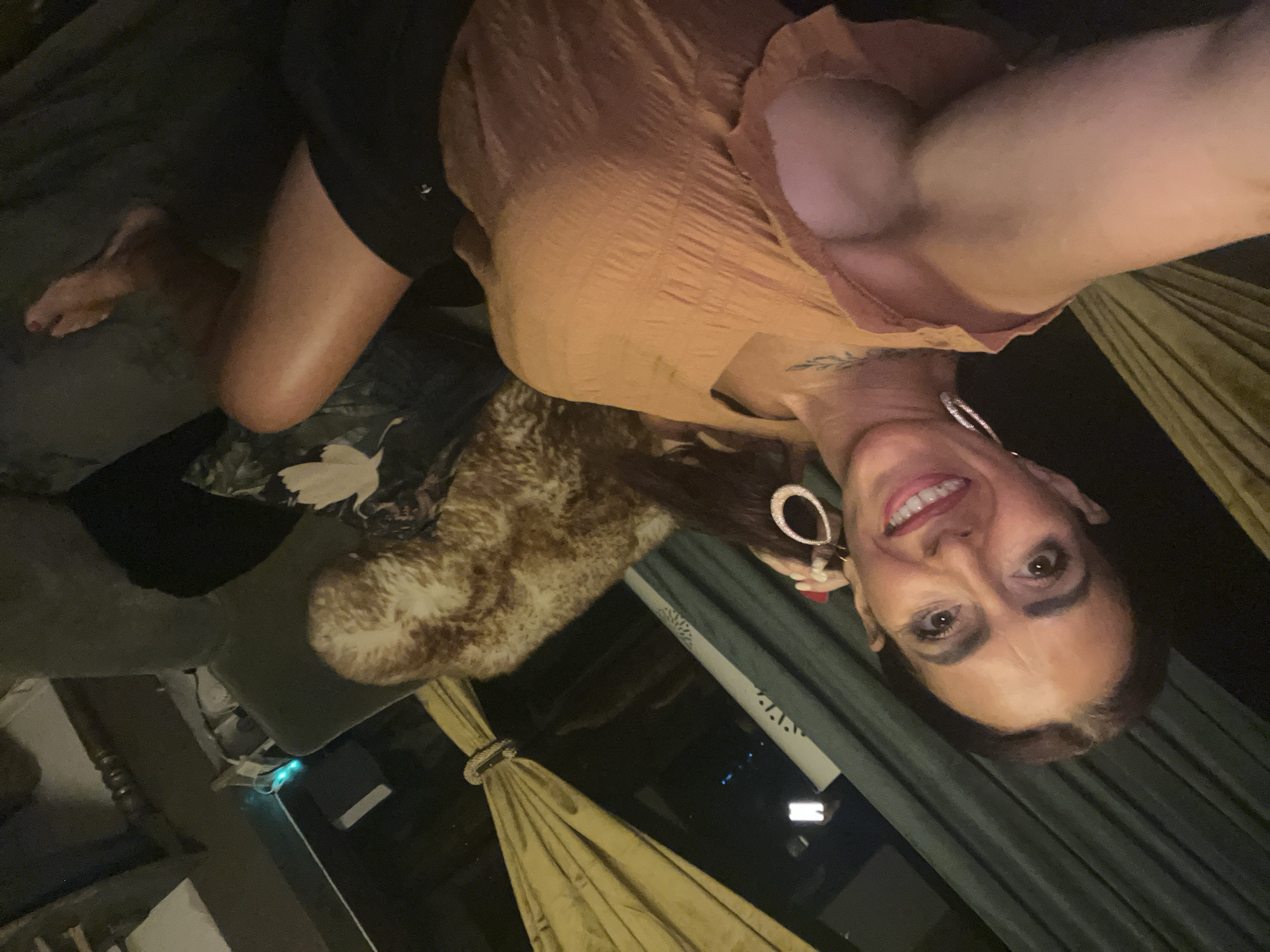 The image shows a woman lying on a sofa, taking a selfie. She is wearing an orange top and black shorts. She has long dark hair and is wearing large hoop earrings. A furry throw pillow is visible behind her. The scene appears to be indoors, likely a living room. There are no visible landmarks or clues to suggest a specific location within Konz, Germany.