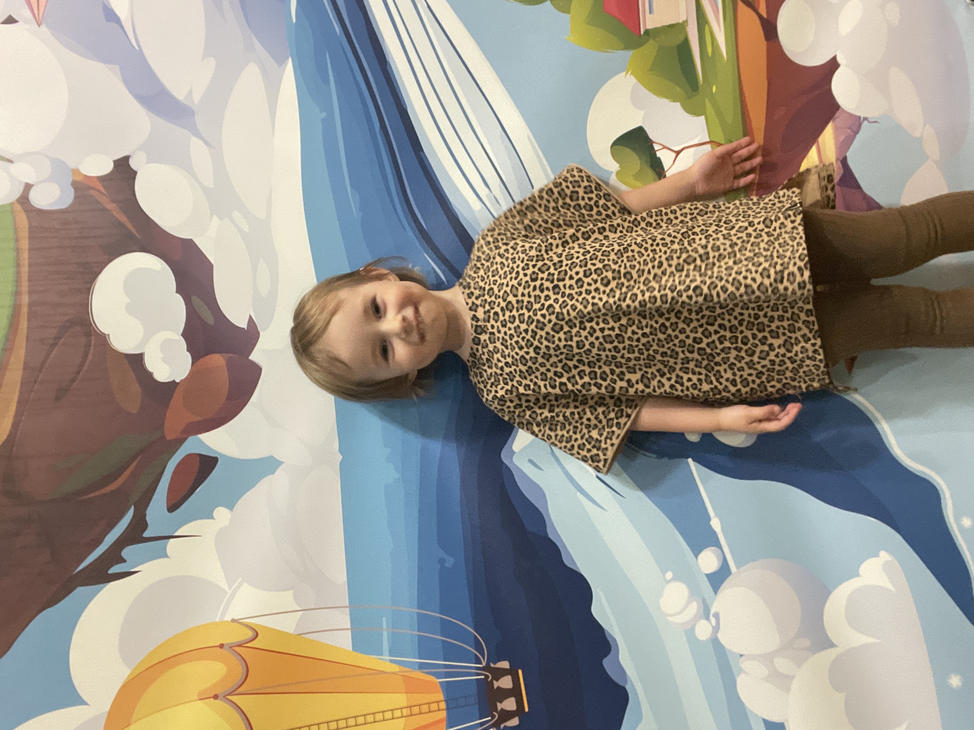A young child with blonde hair, wearing a leopard-print shirt and brown leggings, stands in front of a colorful mural. The mural depicts a fantastical landscape with a yellow hot air balloon, floating islands with trees and houses, blue water, and stylized clouds. The child is smiling and looking at the camera, with their right hand resting on their hip. The lighting suggests the photo was taken indoors during the day. The mural is the dominant feature of the background. There is no information available to specify a location in Vladivostok, Russia.